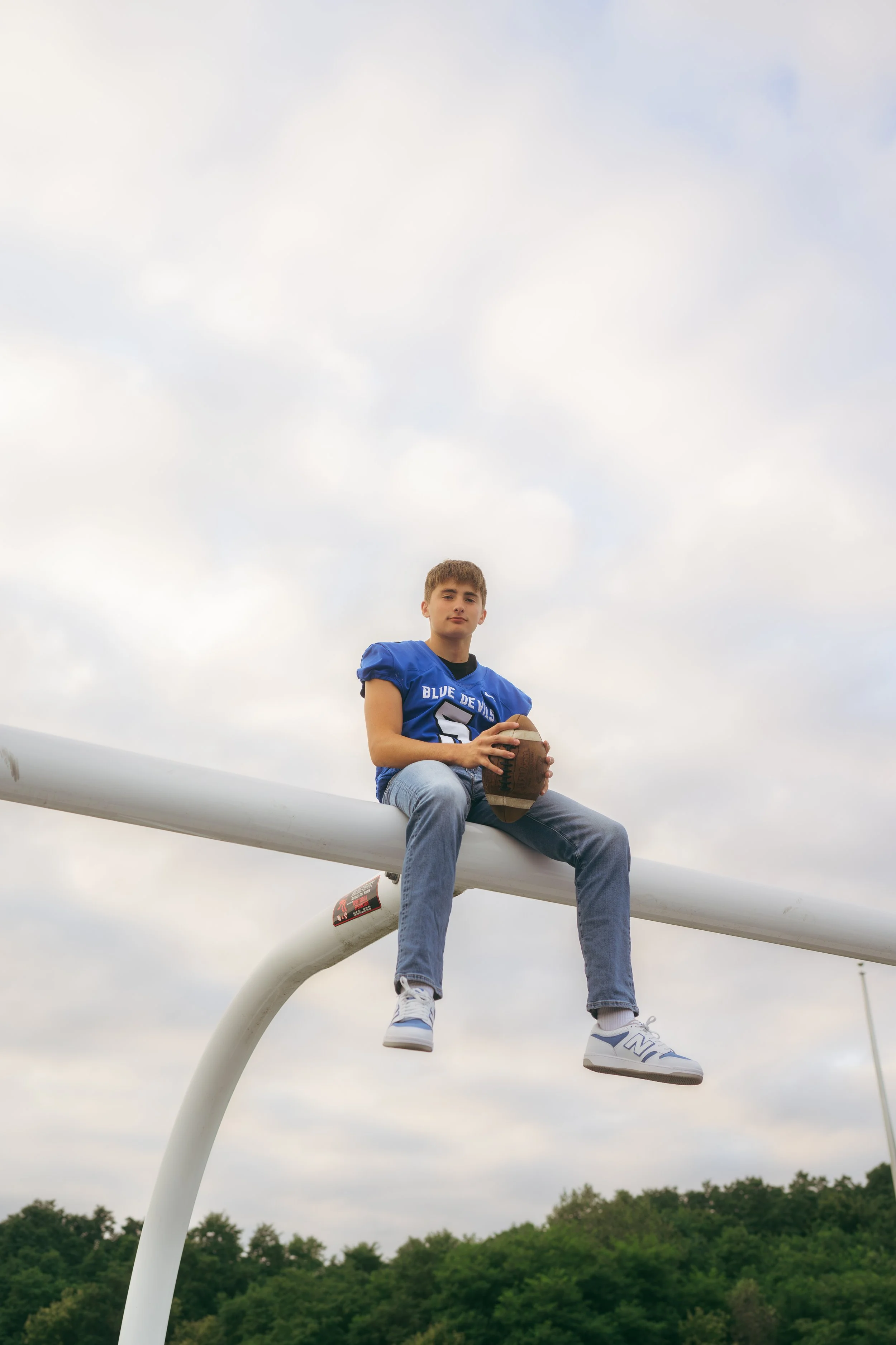 A young man sitting on a football goalpost holding a football, wearing a blue football jersey and jeans, with a cloudy sky and trees in the background.