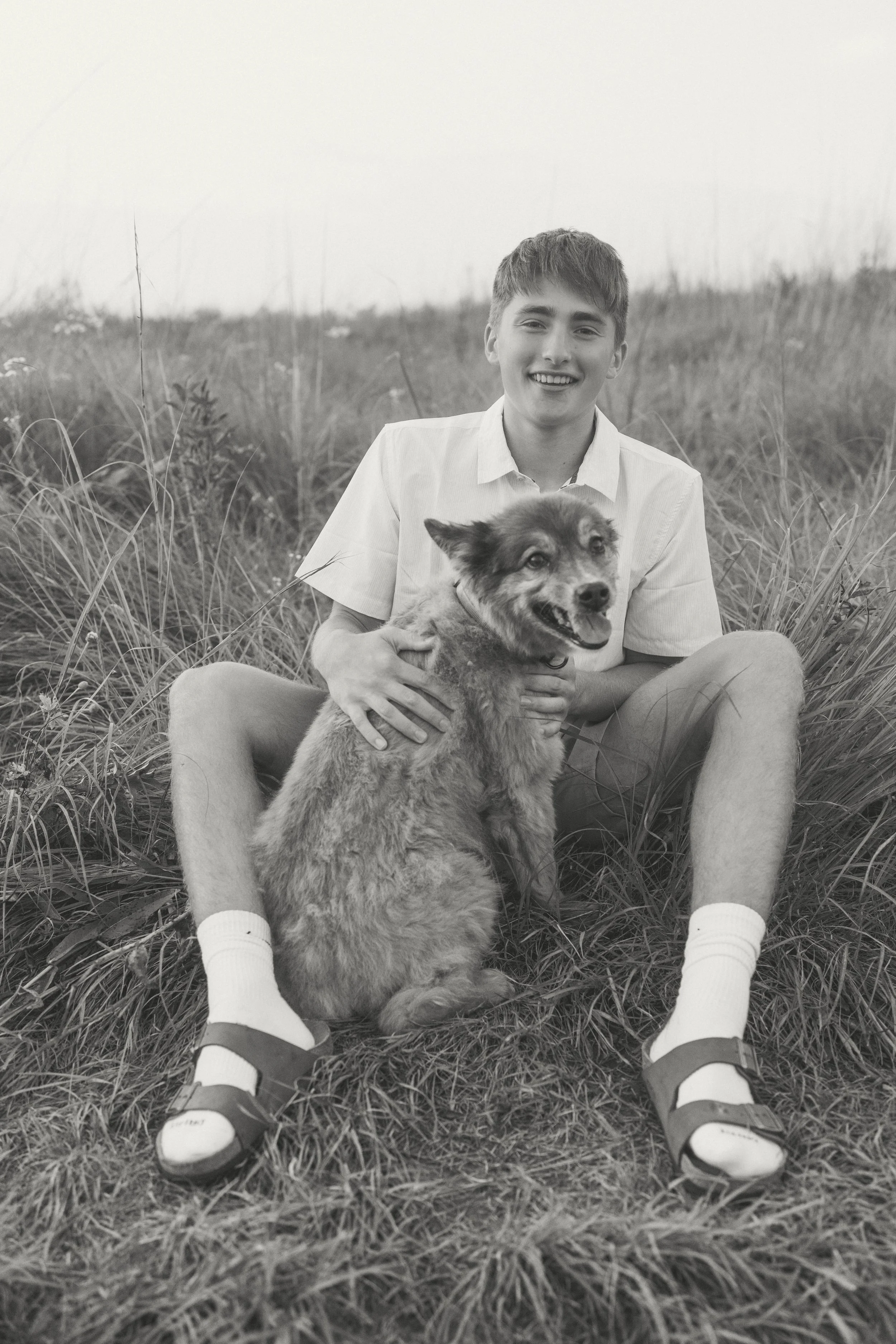 A young man sitting in a grassy field holding a mixed breed dog with a happy expression.