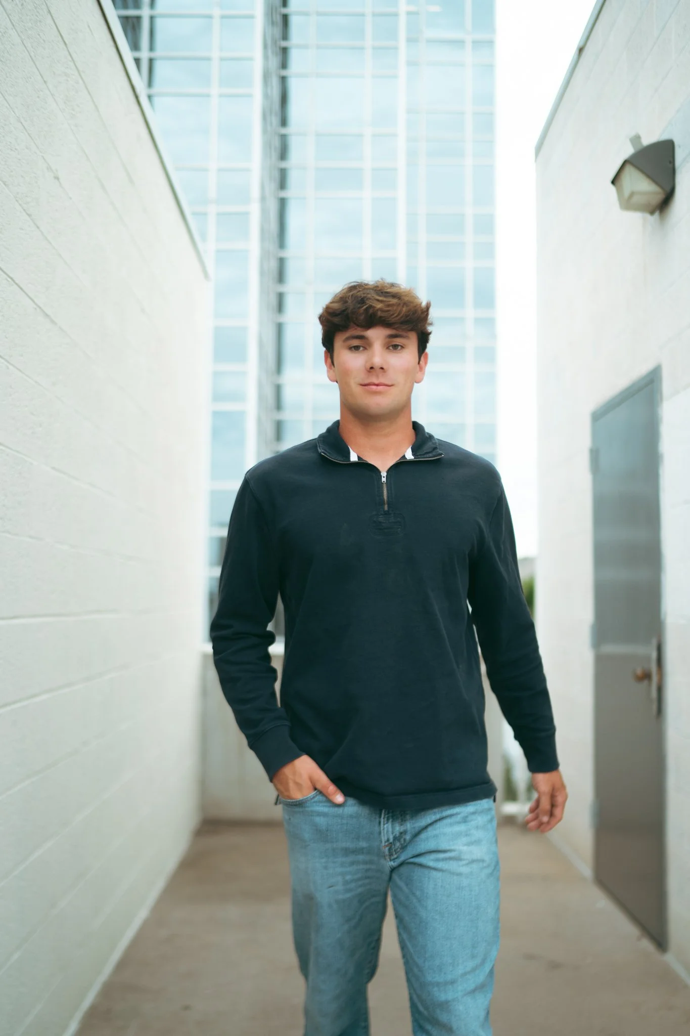 A young man with brown hair wearing a black sweatshirt and light blue jeans walking outdoors in an alleyway with modern glass buildings in the background.