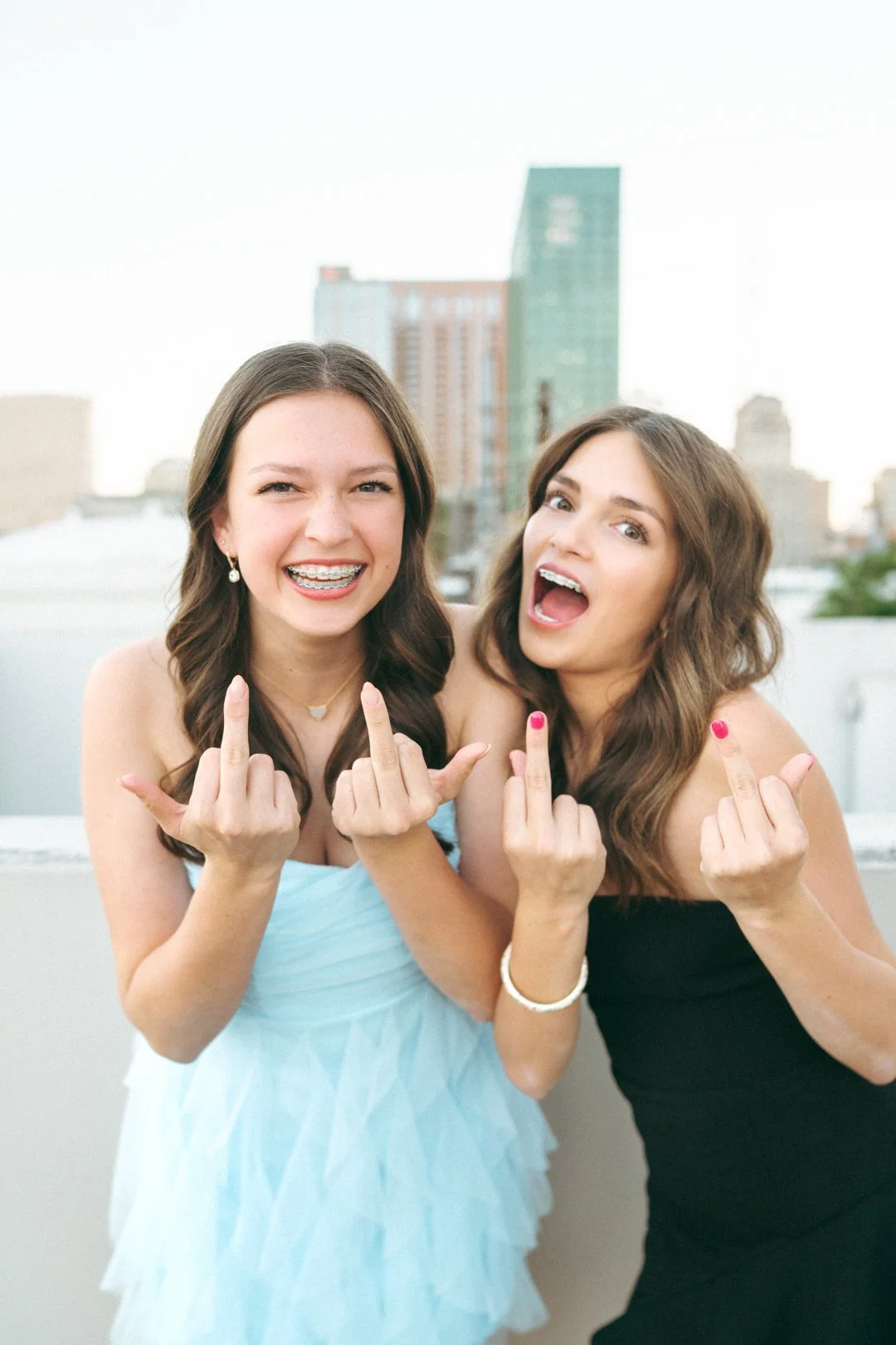 Two young women smiling and making a hand gesture with their middle fingers raised, standing outdoors against a city skyline background.