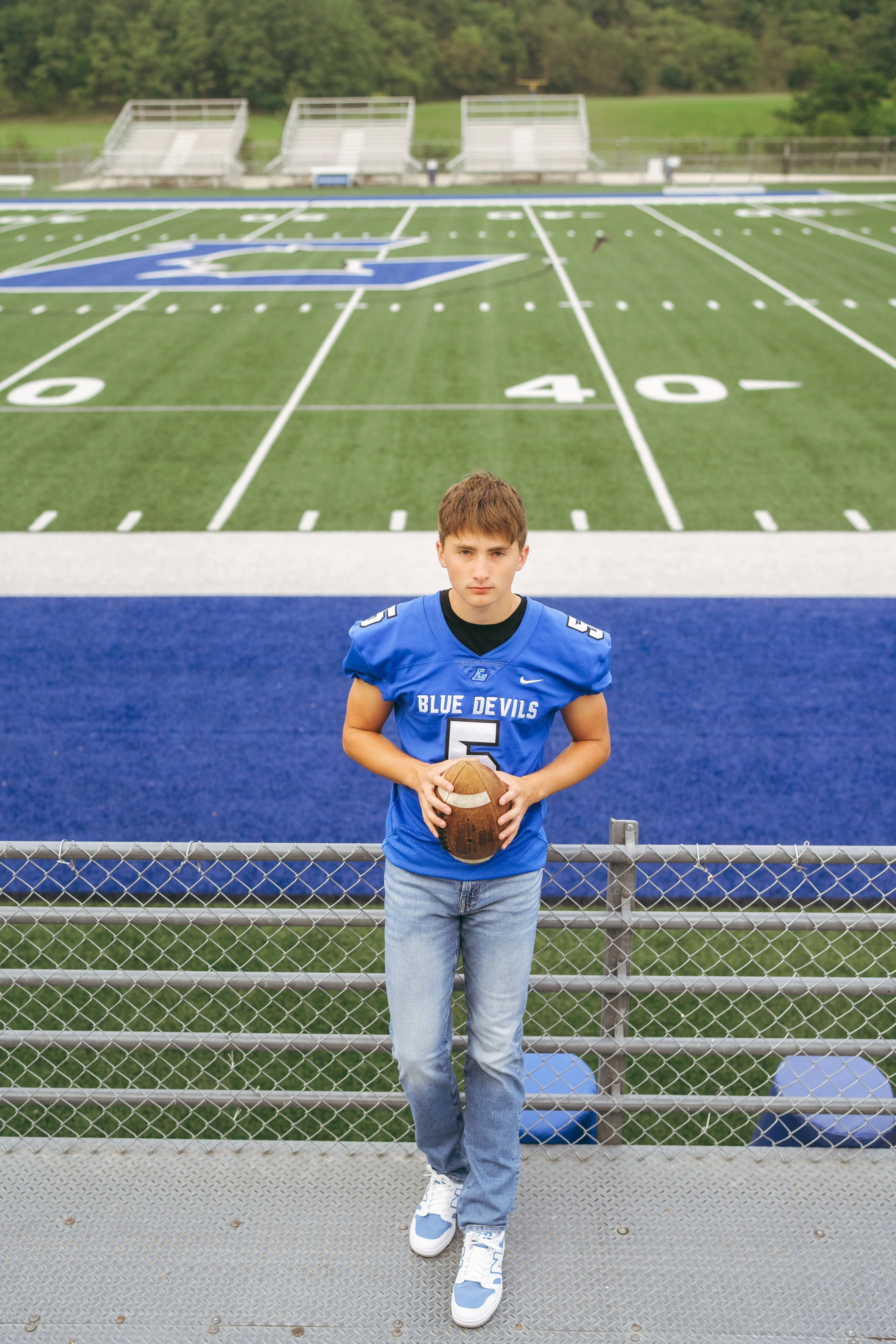 A young boy wearing a blue football jersey with the number 5 and 'Blue Devils' written on it, holding a football, standing on a track near a football field with American football markings.