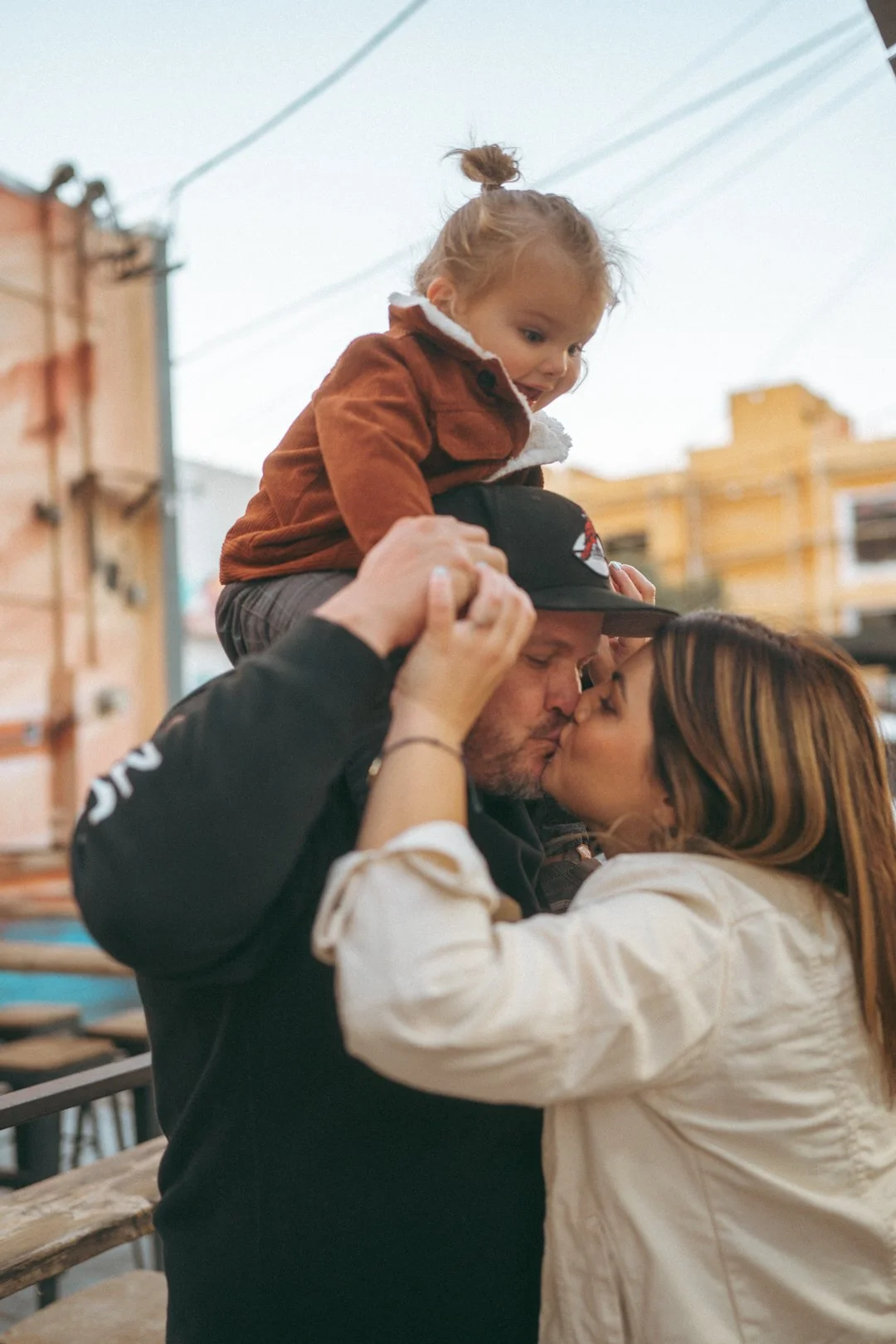 Family of three sharing a kiss outdoors on a city street, with a young girl sitting on a man's shoulders, and a woman and man kissing in front of each other.