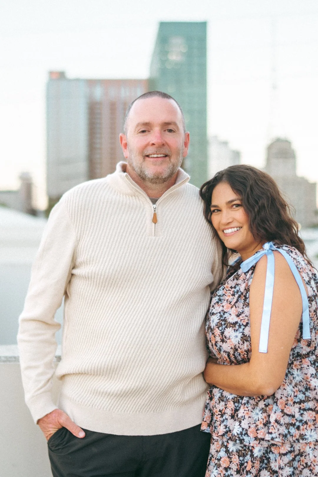 A man and woman smiling outdoors against a city skyline with tall buildings in the background.