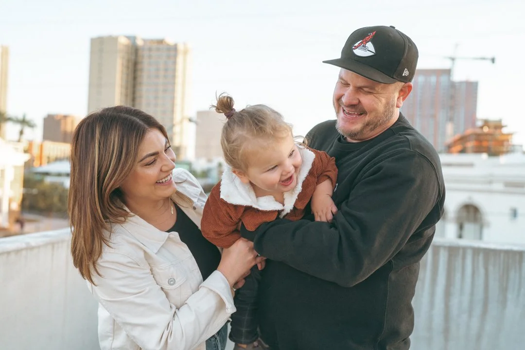 Family of three on rooftop with city buildings in background, smiling and laughing together.
