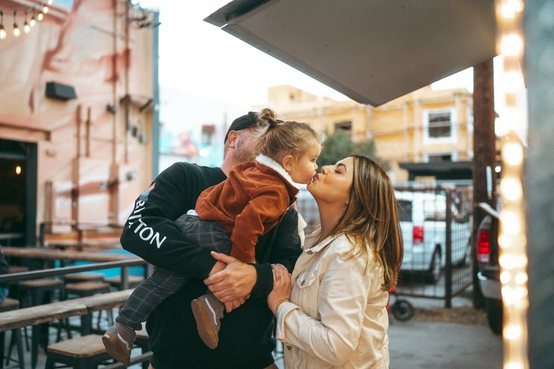 A family sharing a kiss in an outdoor setting, with a child leaning in to kiss a woman, while a man holds the child.