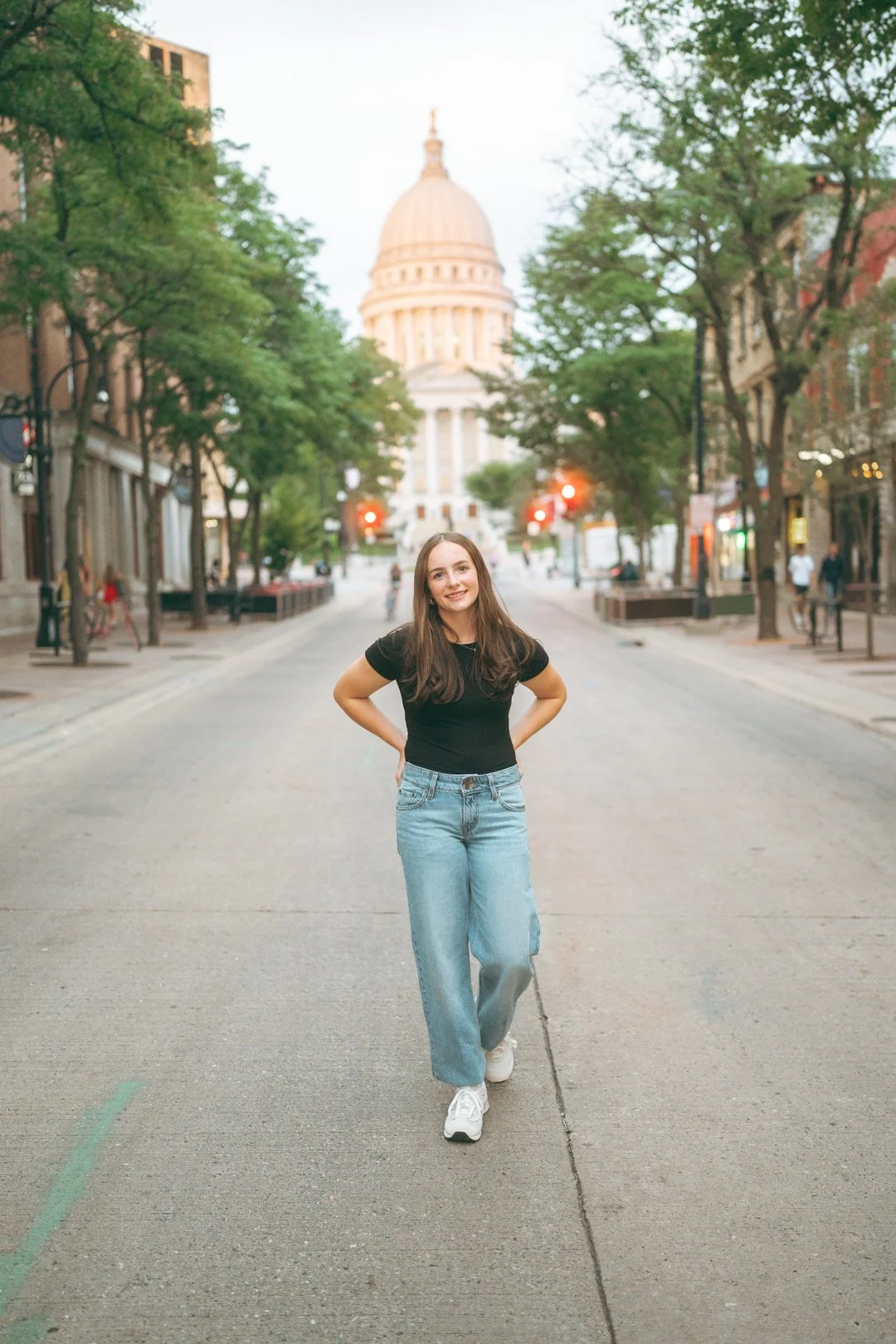 A woman with long brown hair, wearing a black t-shirt, light blue jeans, and white sneakers, walking down a city street with the state capitol building in the background, surrounded by trees and buildings.