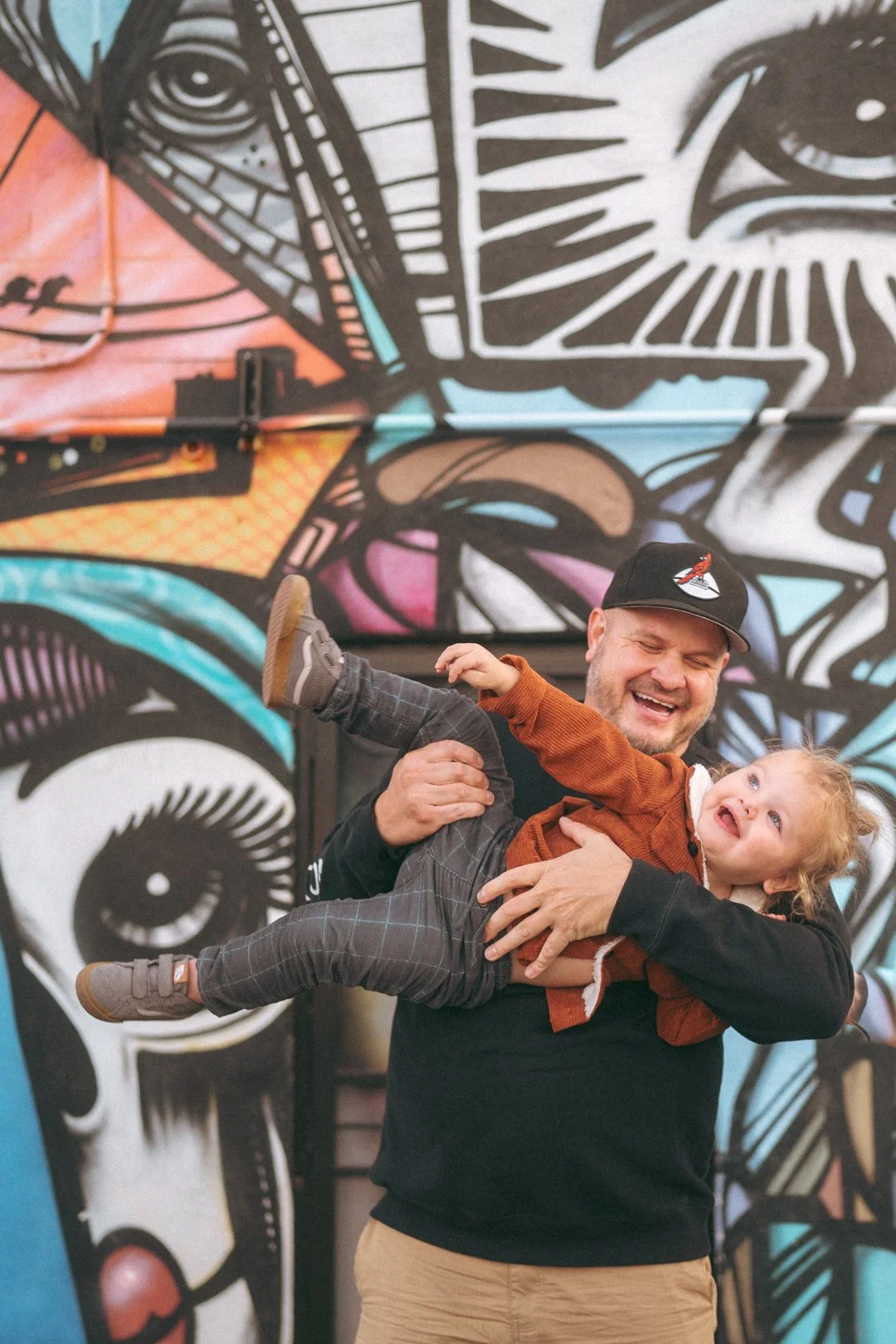A man with a cap smiling and holding a young girl in front of a colorful graffiti mural, with the girl looking up and smiling.
