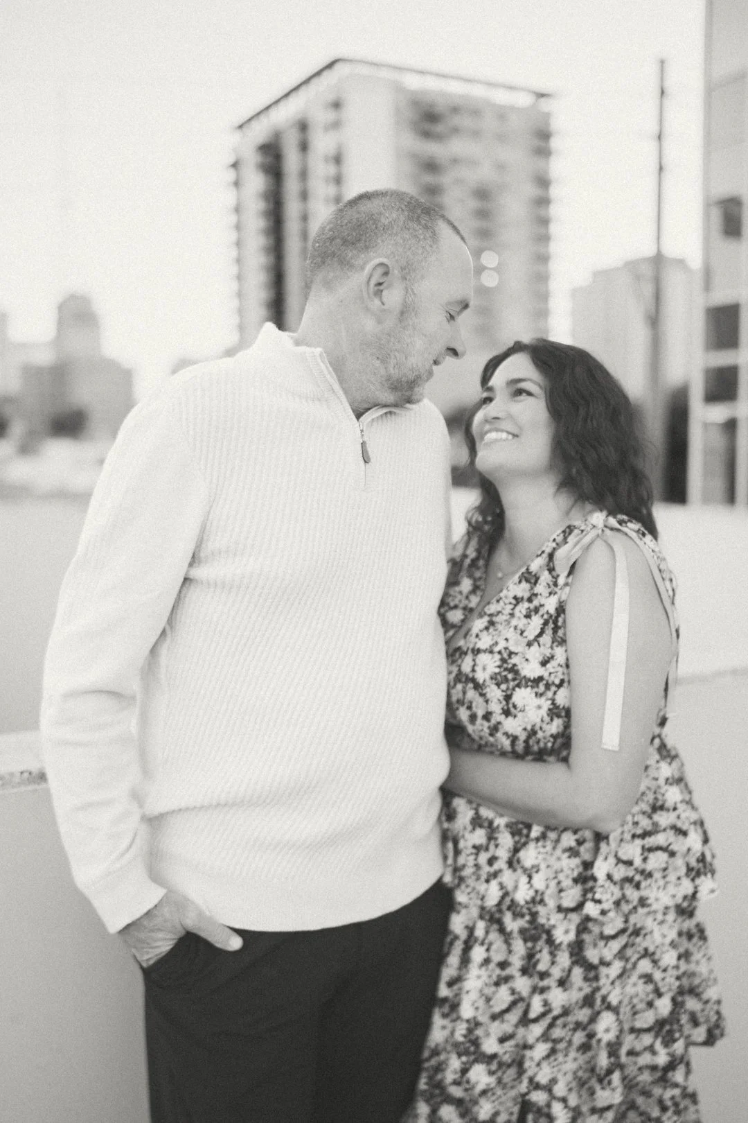 A black-and-white photo of a man and woman standing close together outdoors in an urban setting, smiling at each other.