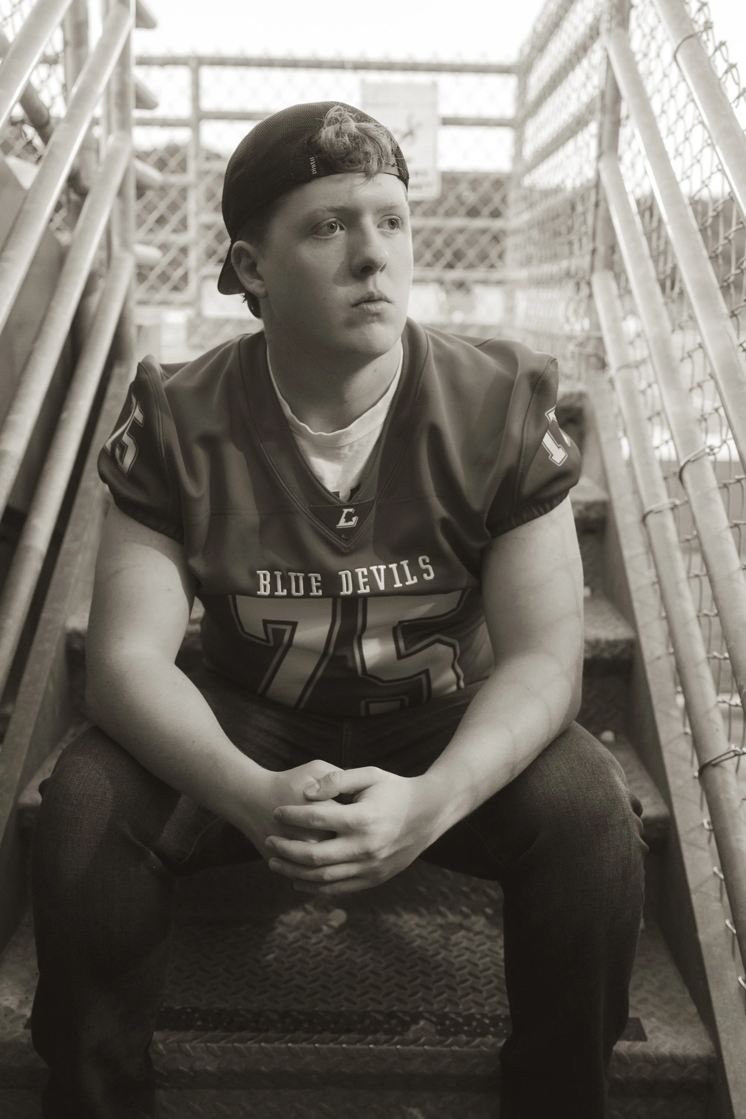 A young male football player sitting on stadium bleachers, wearing a football jersey with 'Blue Devils' on it, a baseball cap backward, and looking contemplative.