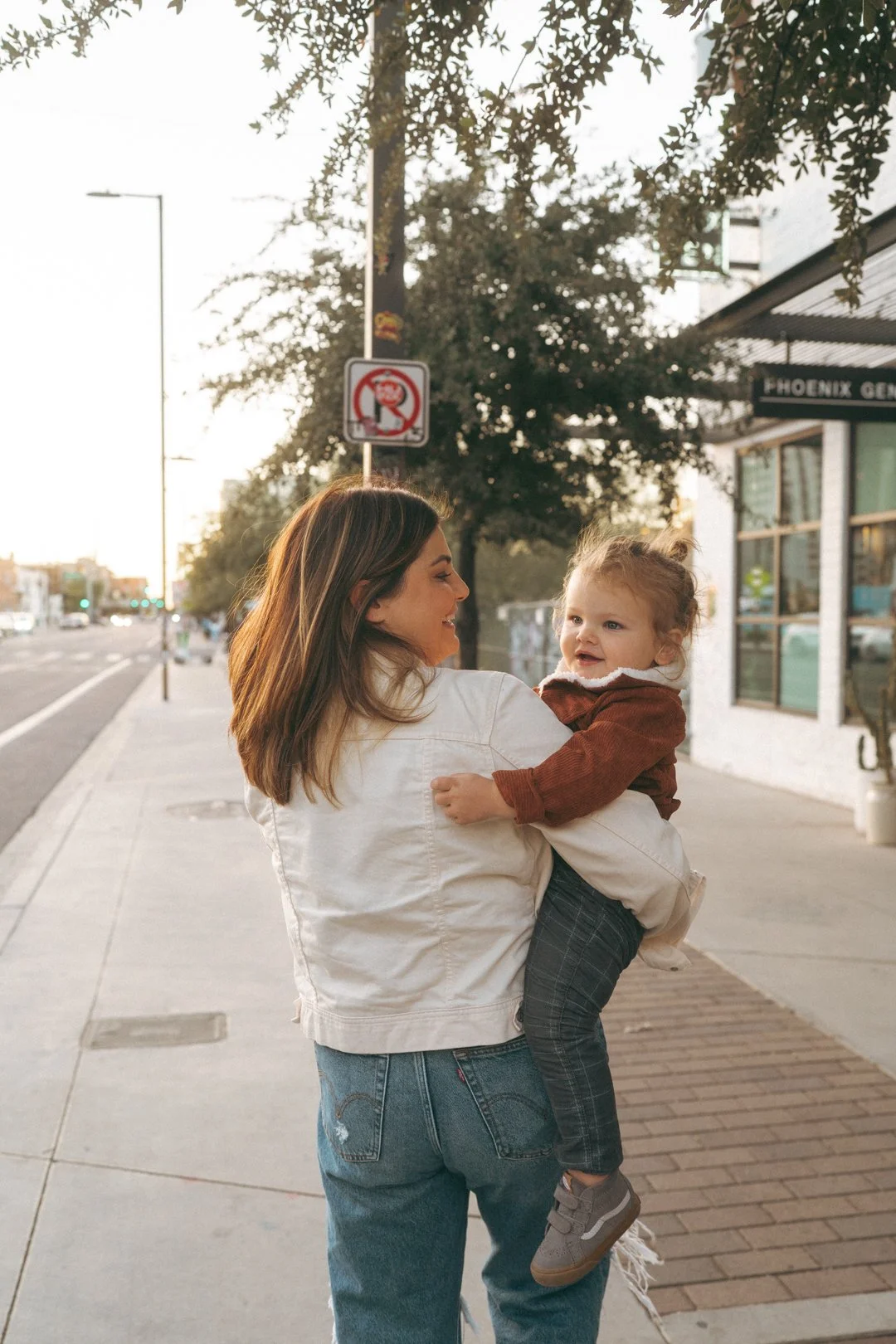 A woman holding a young girl in a casual outdoor setting on a sidewalk during daylight. The woman is wearing a white jacket and jeans, while the girl is dressed in a brown jacket and checkered pants. There are trees, a street, and storefronts in the 