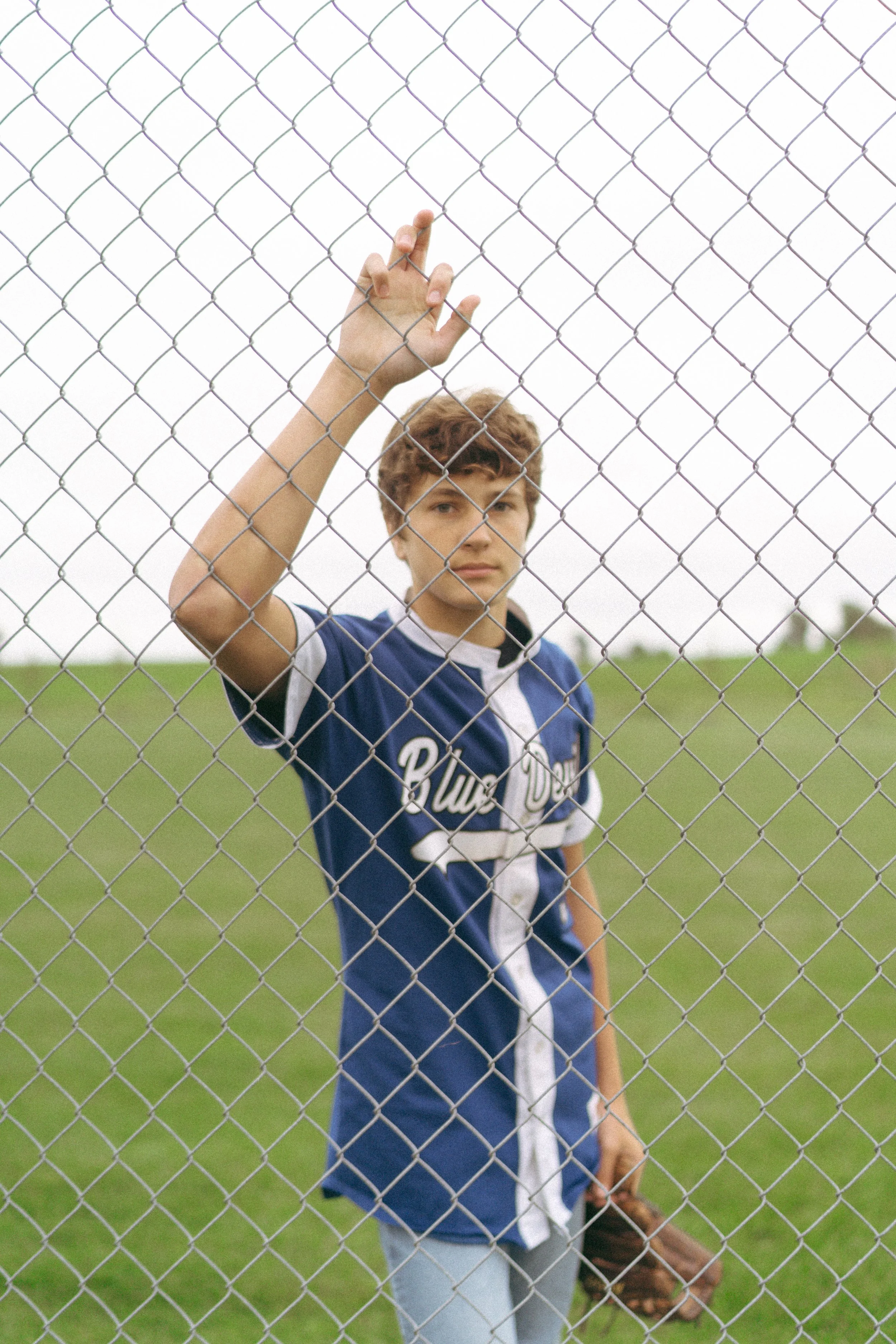 Teen boy in a blue baseball jersey and jeans standing behind a chain-link fence on a grassy field, holding a baseball glove in his left hand, and raising his right hand with fingers touching the fence.