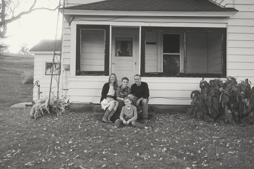 Family sitting on steps in front of a house, with front porch missing two sections of railing, and a lawn with fallen leaves.