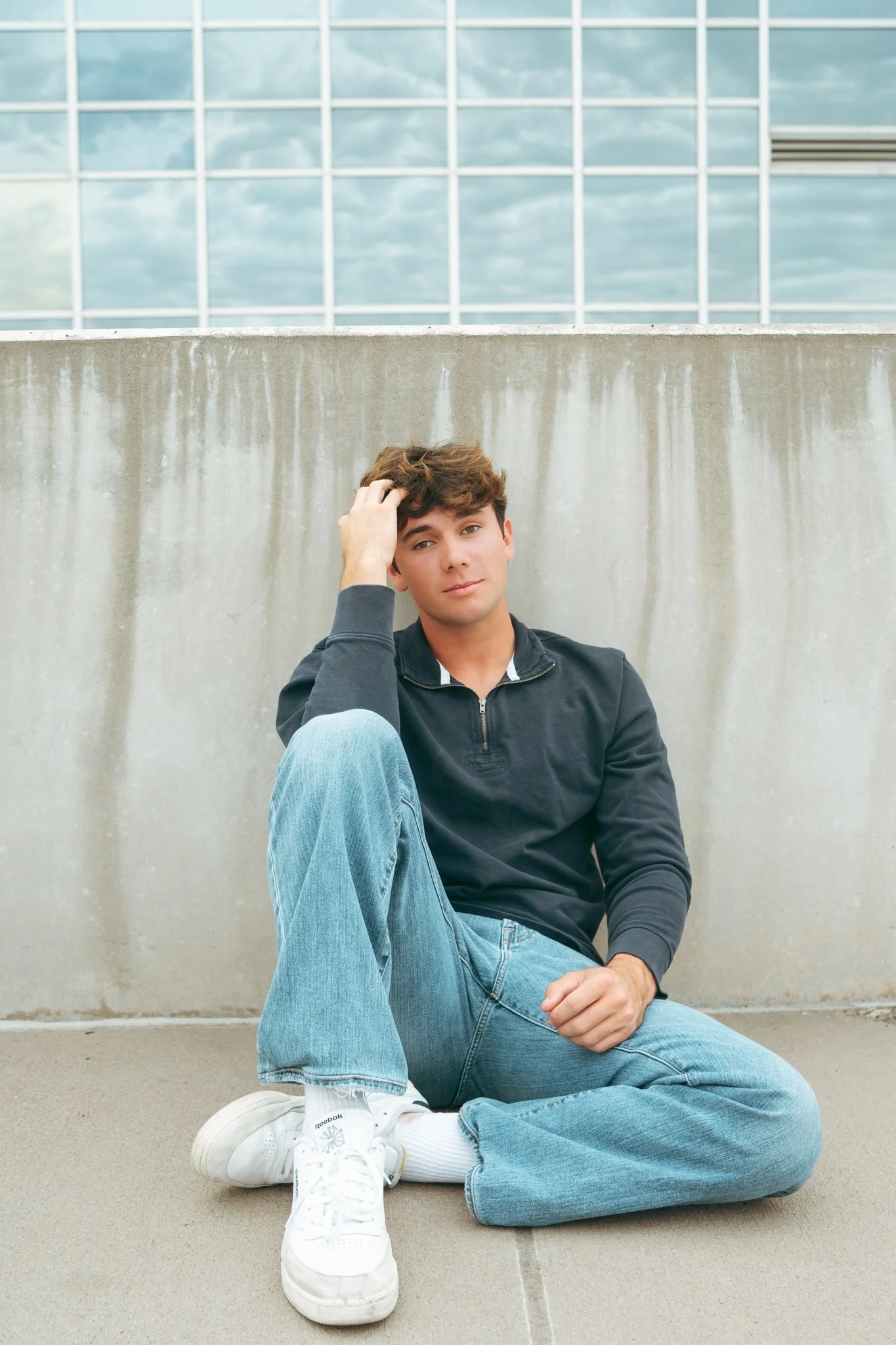 A young man sitting on the ground in front of a concrete wall, with a modern glass building behind him. He is wearing a black quarter-zip sweatshirt, light blue jeans, and white sneakers. He has brown hair and is looking at the camera with a neutral 