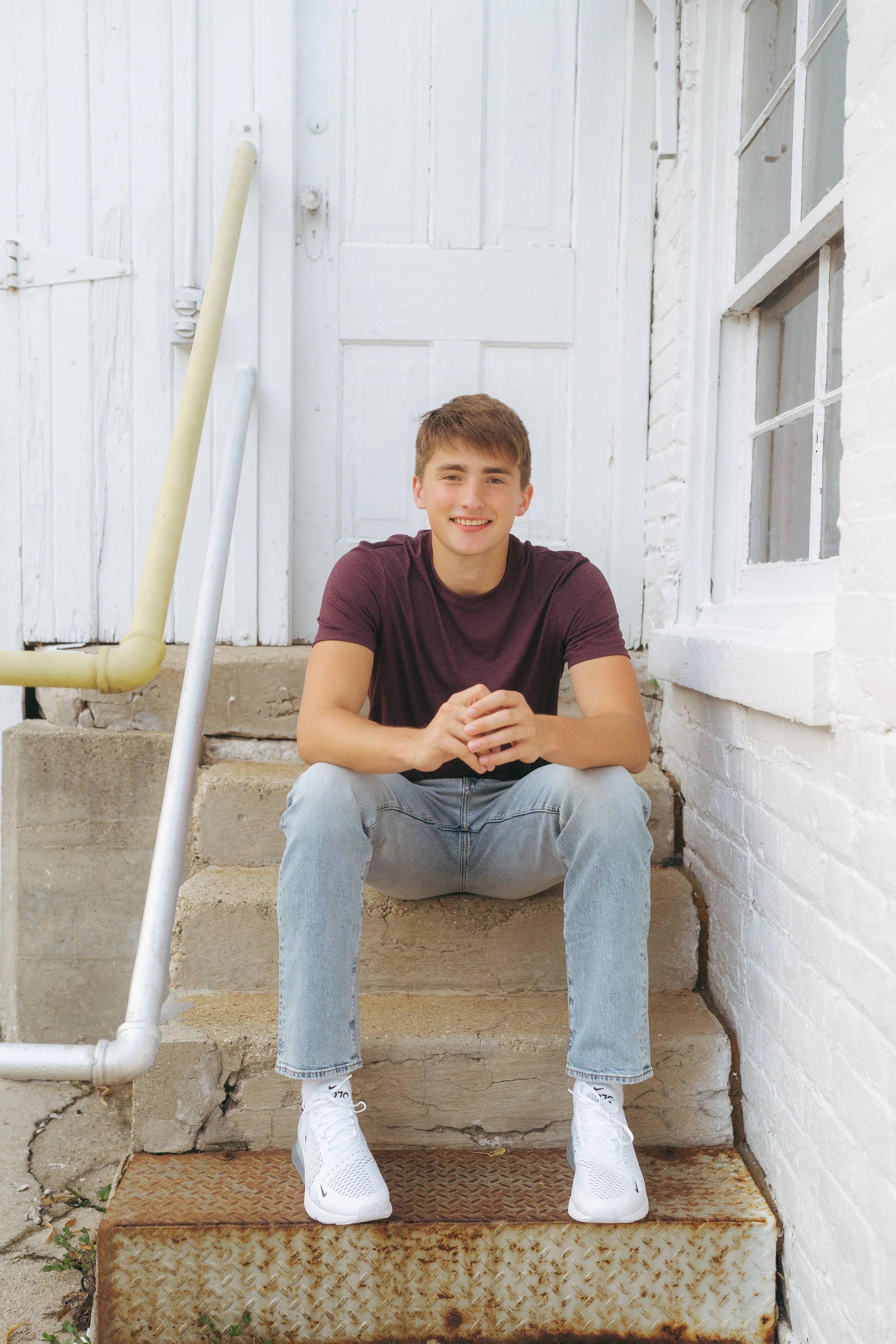 A young man with short brown hair, wearing a maroon t-shirt, light blue jeans, and white sneakers, sitting on the outdoor concrete steps of a white brick building, smiling at the camera.