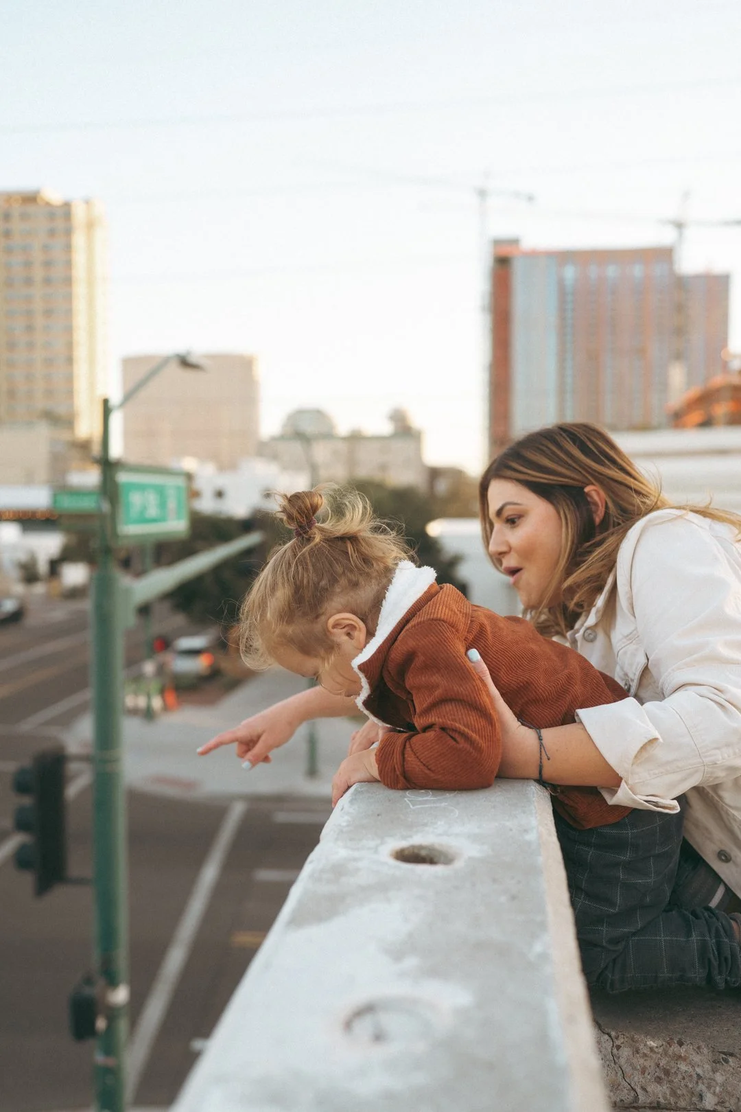 Woman and young girl leaning over a concrete ledge, looking down at a city street below, pointing at something in the distance.