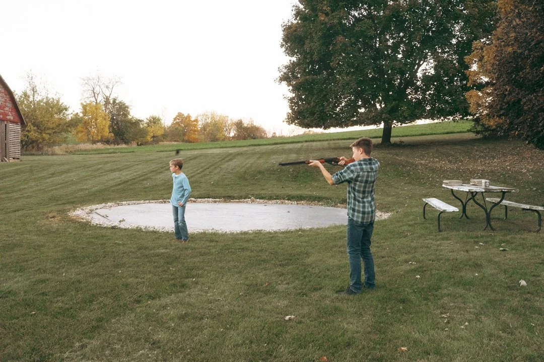 Two boys in a grassy yard, with one boy aiming a rifle and the other standing nearby. The background features a large tree, a small pond or sandpit, and a red barn on the left side.