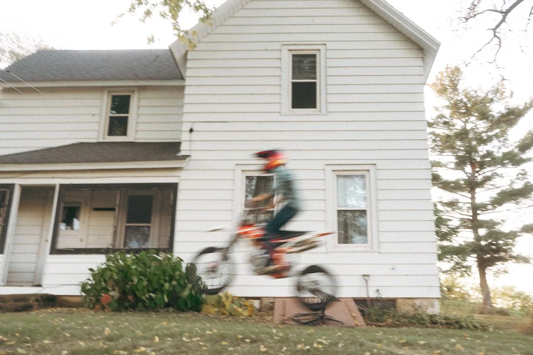 A person riding a dirt bike in front of a white house with a gable roof and multiple windows, slightly blurred from motion.