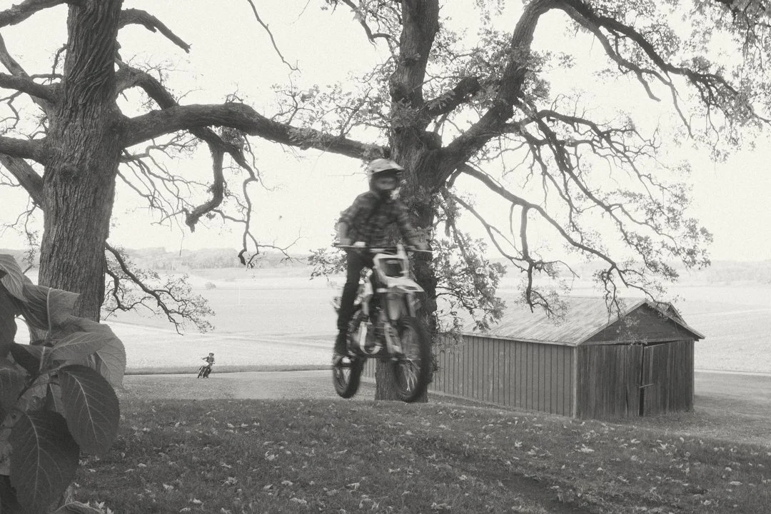 A person riding a BMX bike jumps off the ground under a large leafless tree in a rural area with a barn in the background and open fields.