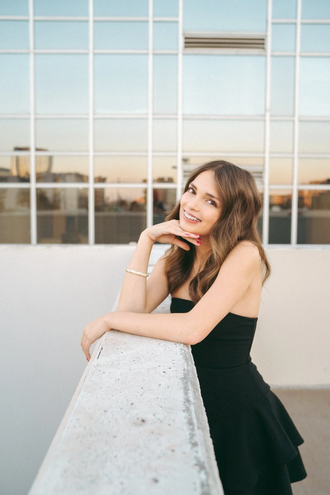 A young woman with wavy brown hair in a strapless black dress, smiling and resting her chin on her hand, standing next to a white concrete ledge with a cityscape and grid-like windows in the background during sunset.