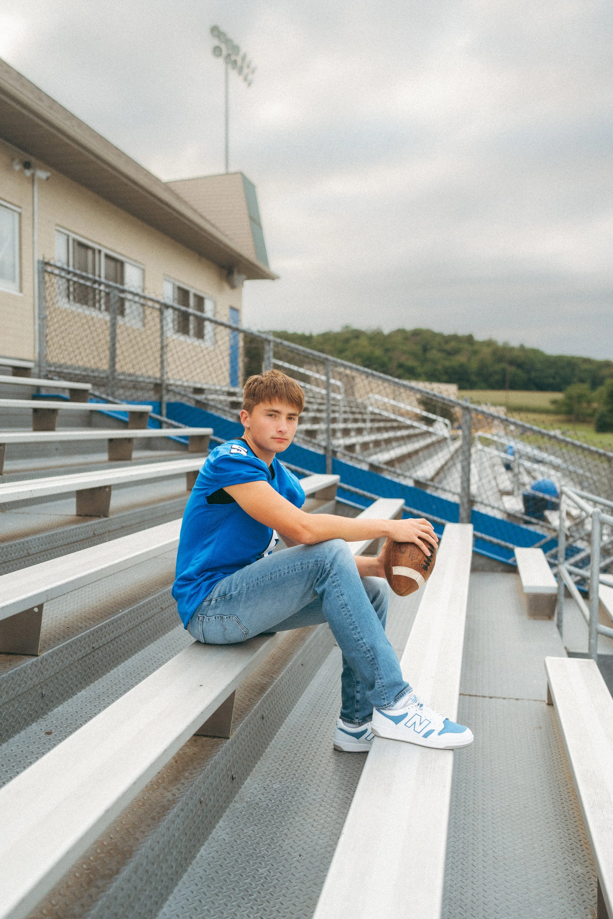 A teenage boy sitting on bleachers in a football stadium, dressed in a blue sports jersey, jeans, and white sneakers, holding a football.