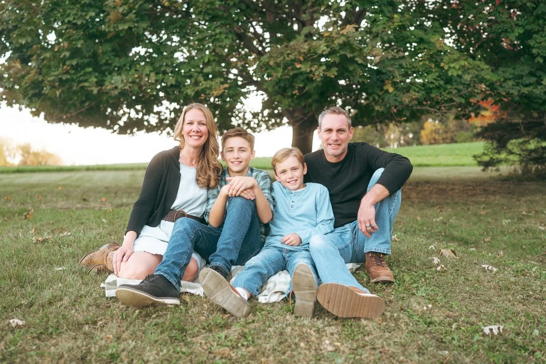 A family of four sitting on grass in a park under a large leafy tree, smiling at the camera.