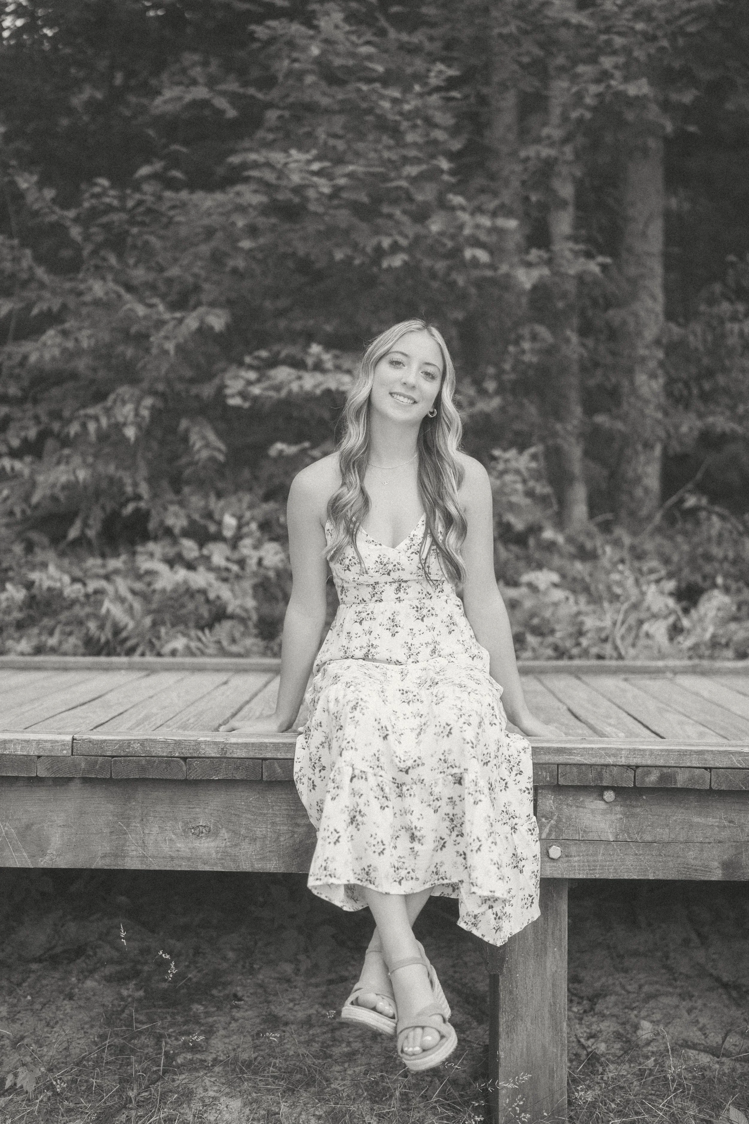 A young woman with long wavy hair, wearing a floral dress and sandals, sitting on a wooden platform outdoors with a background of trees and foliage.