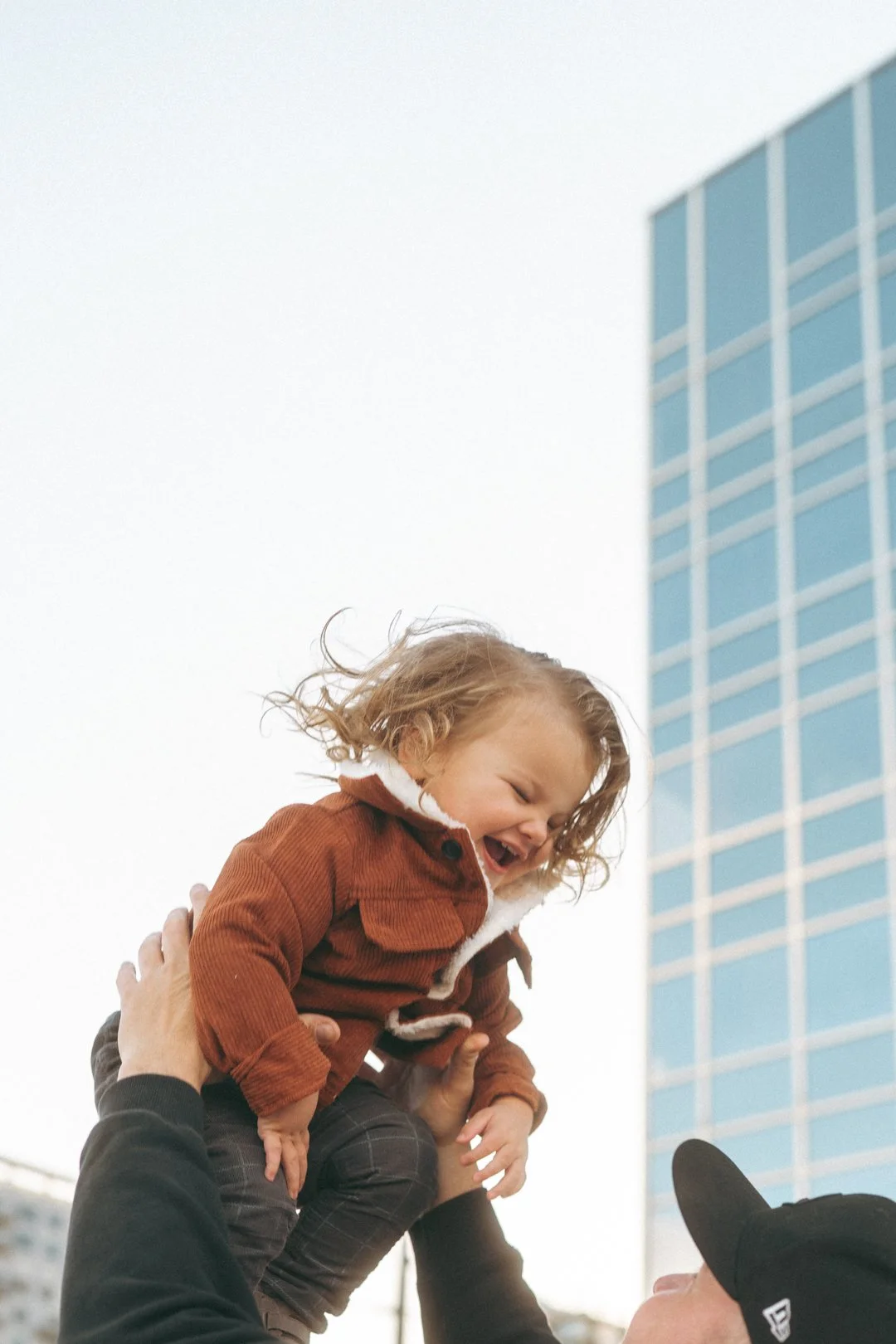 A young girl with curly hair, wearing a brown jacket, being lifted by multiple hands, smiling and laughing outdoors with a tall blue glass building in the background.