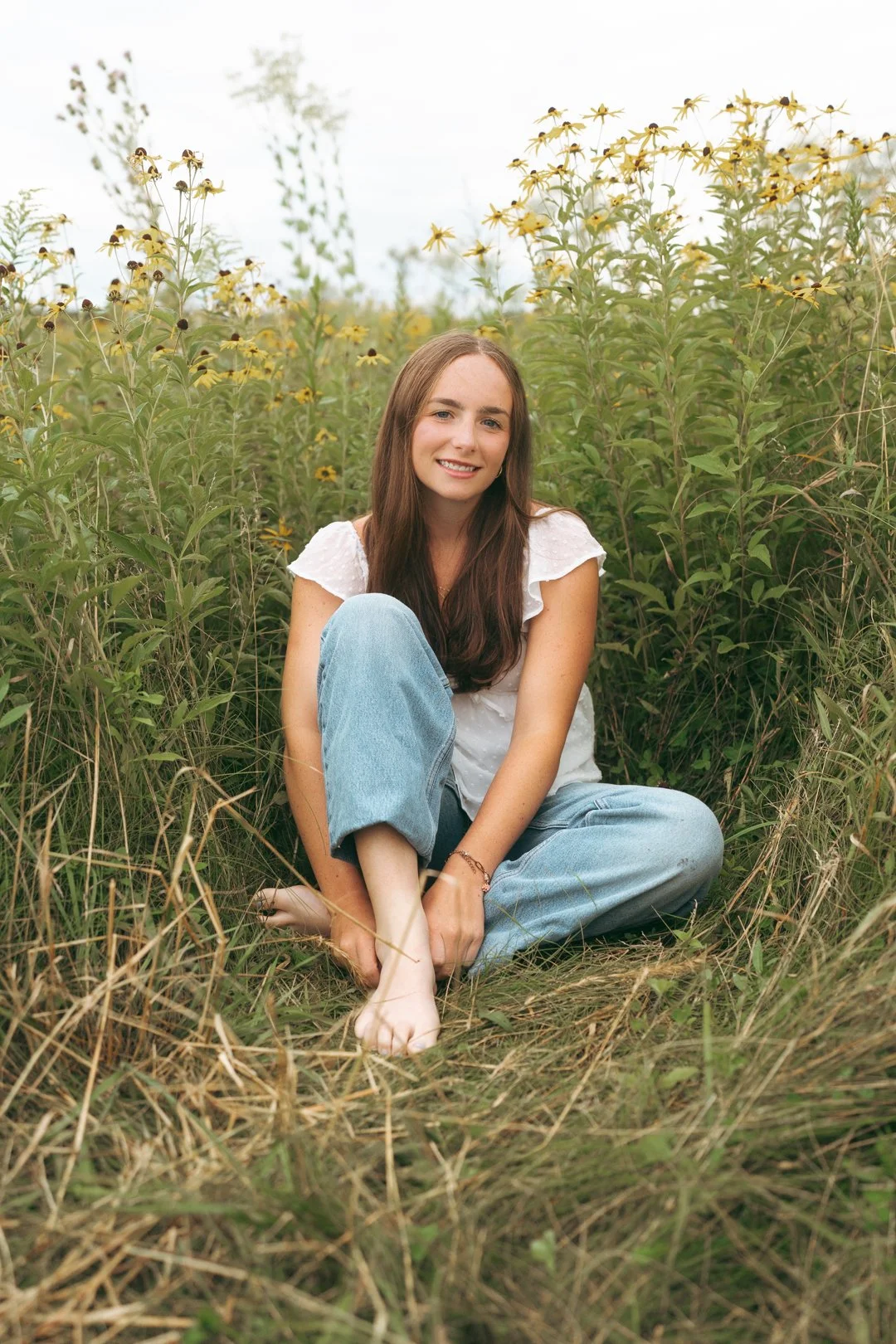 A young woman sitting in a field of yellow coneflowers, smiling at the camera, wearing a white top and light blue jeans.