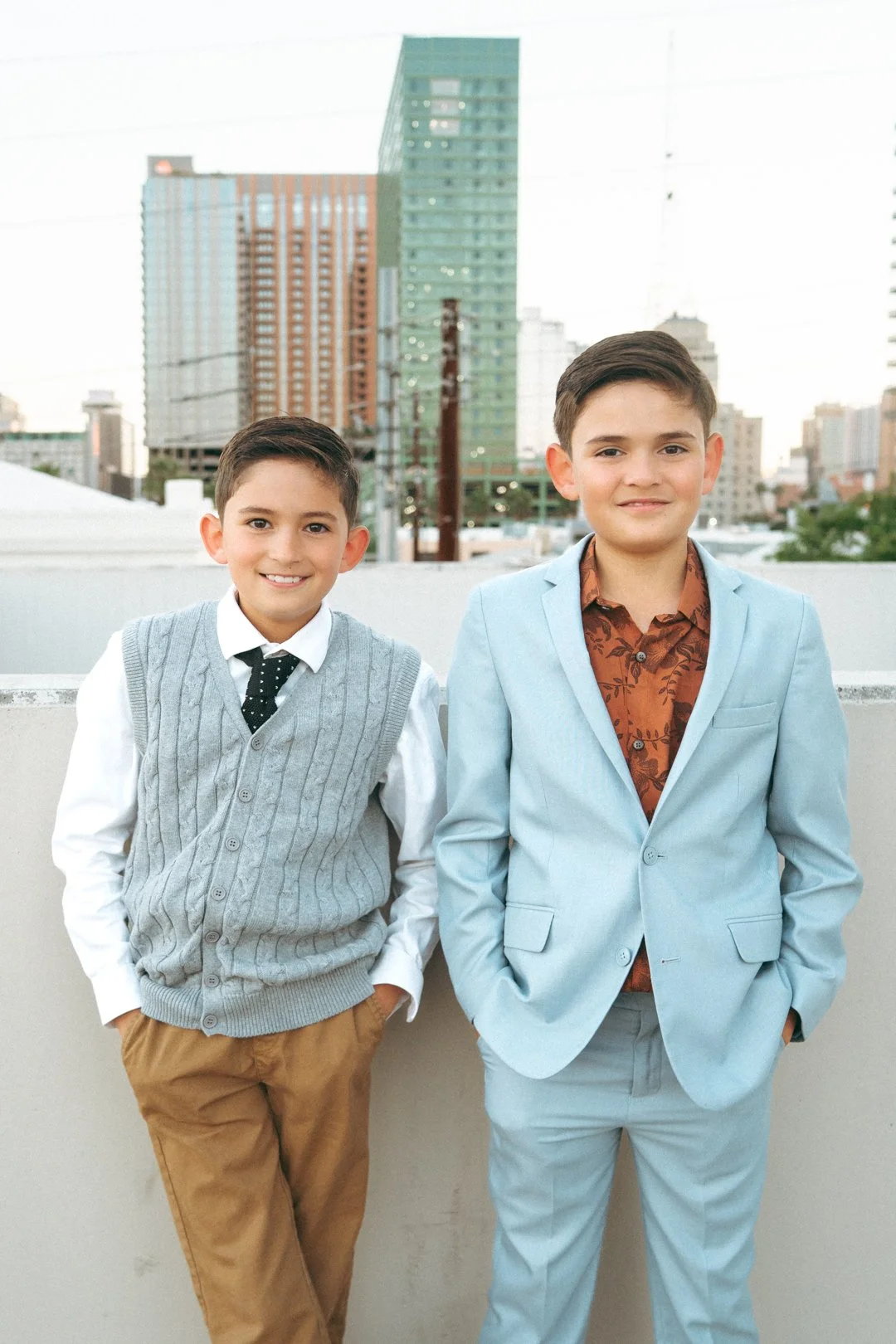 Two young boys in formal attire stand on a rooftop with a cityscape of tall buildings in the background. The boy on the left wears a gray vest, white shirt, and polka dot tie, and the boy on the right wears a light blue suit with a brown patterned sh
