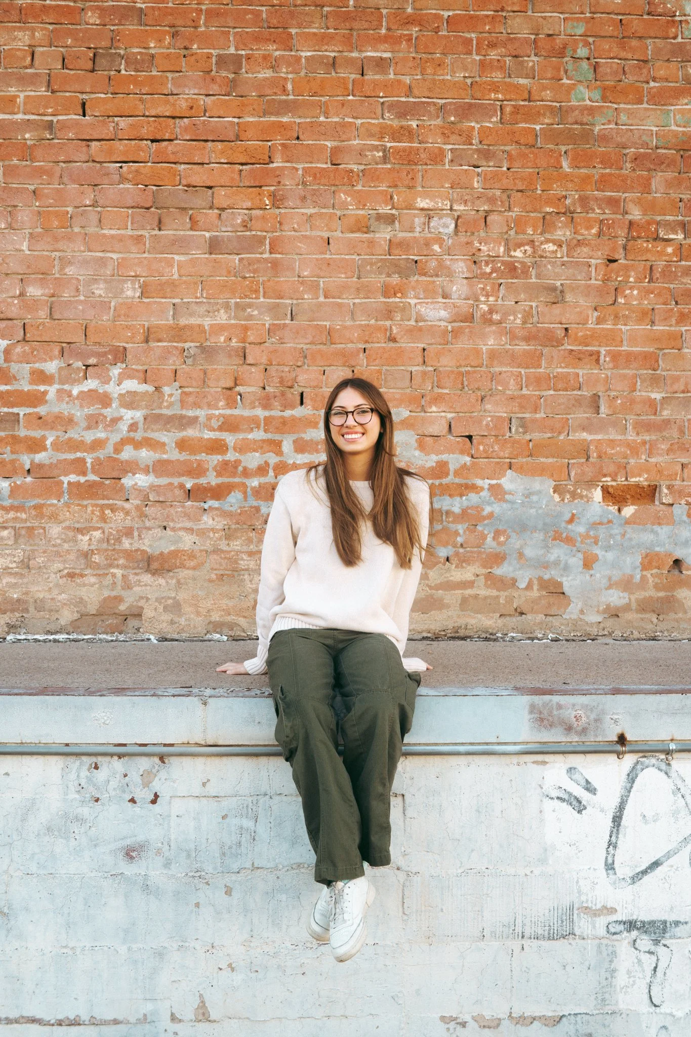 Young woman with glasses sitting on a ledge against a weathered brick wall, smiling at the camera.