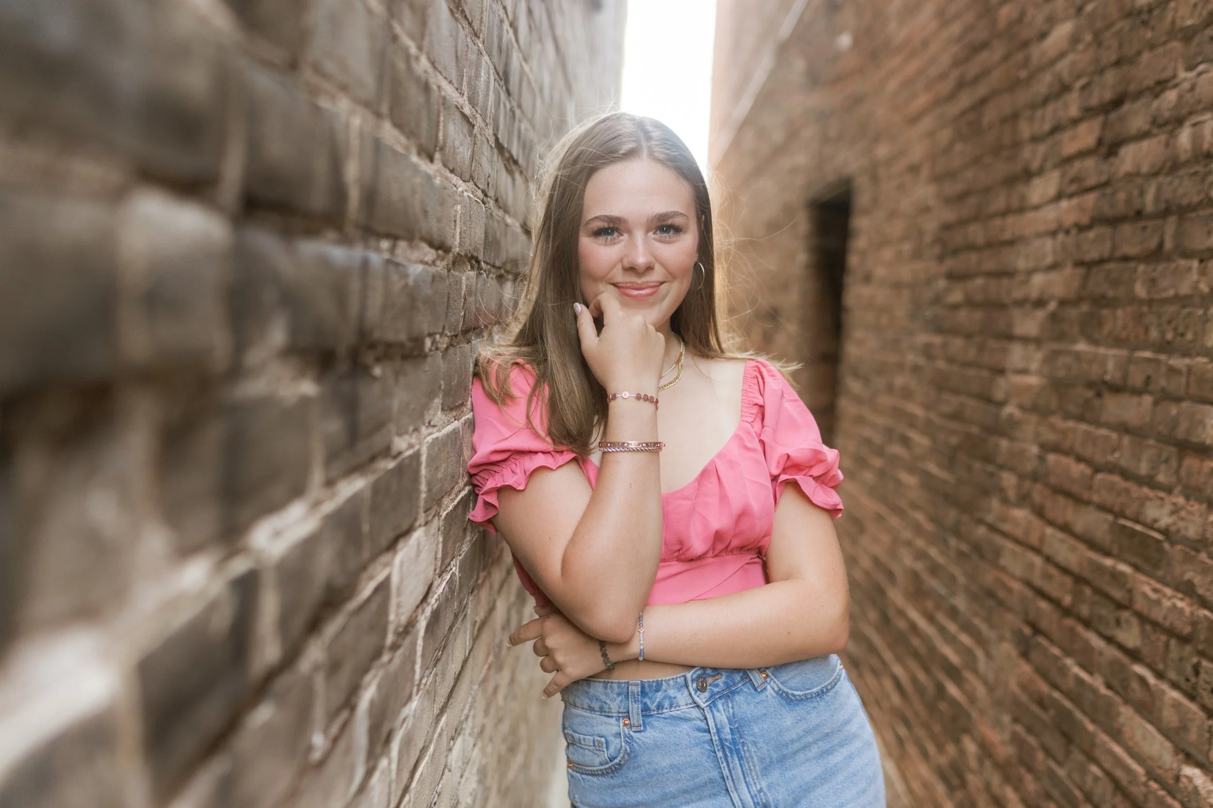 Young woman with long brown hair wearing a pink top and blue jeans, standing between two brick walls, smiling at the camera.