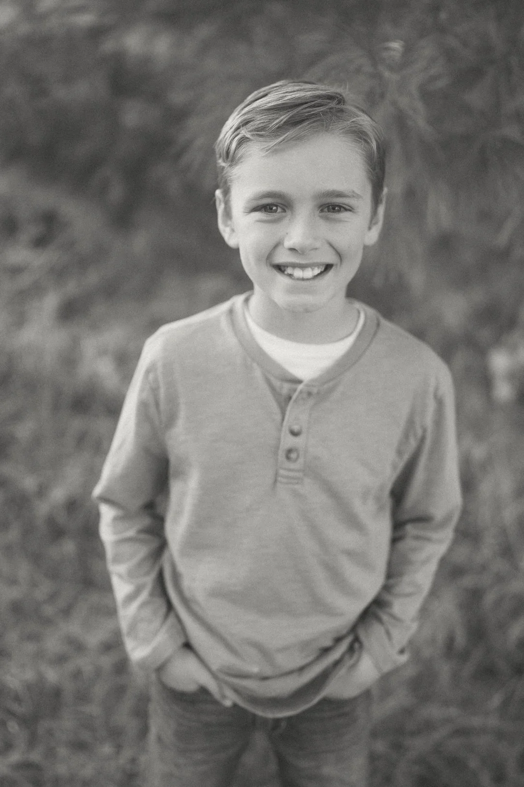 A young boy smiling outdoors with hands in his pockets, wearing a long-sleeve shirt and standing in front of blurred natural background.