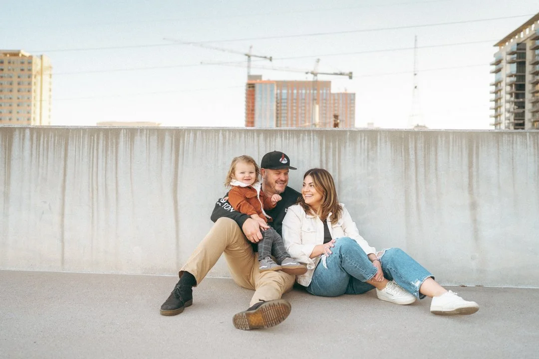A family of three sitting on the ground outdoors in an urban setting, smiling and enjoying each other's company. The father is holding their young daughter, and the mother is sitting beside them.