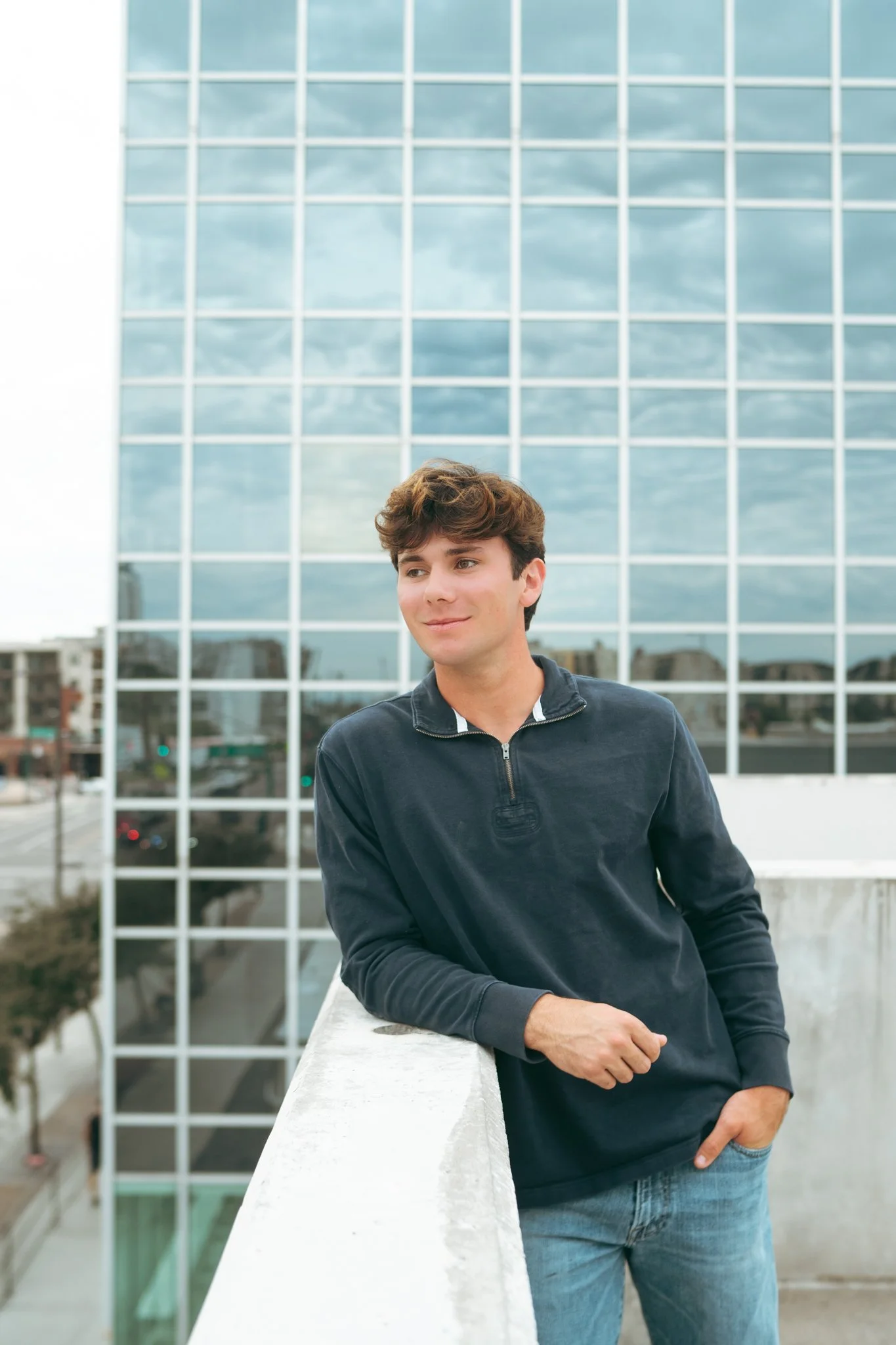 A young man in a navy blue sweatshirt and jeans leaning on a white railing on a rooftop, with a modern glass office building reflecting the cloudy sky in the background.
