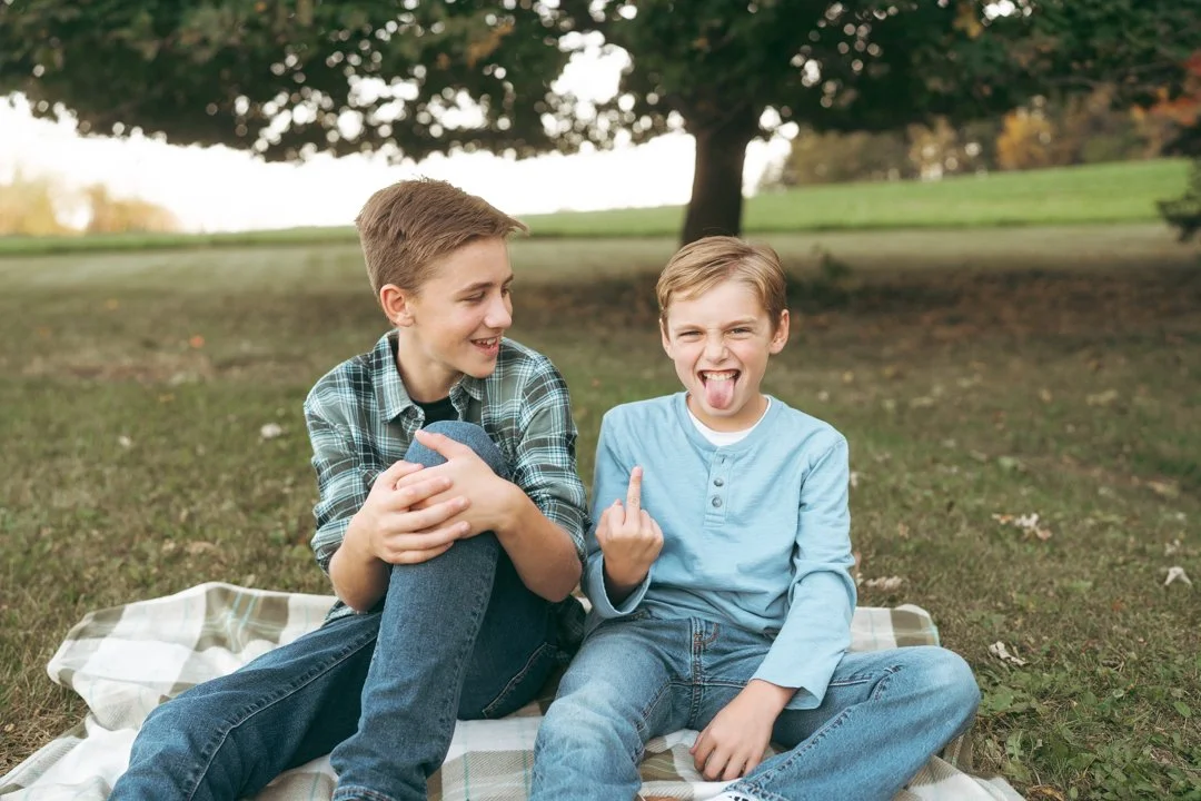 Two boys sitting on a plaid blanket outdoors under a tree in a park, smiling and making silly faces at each other.