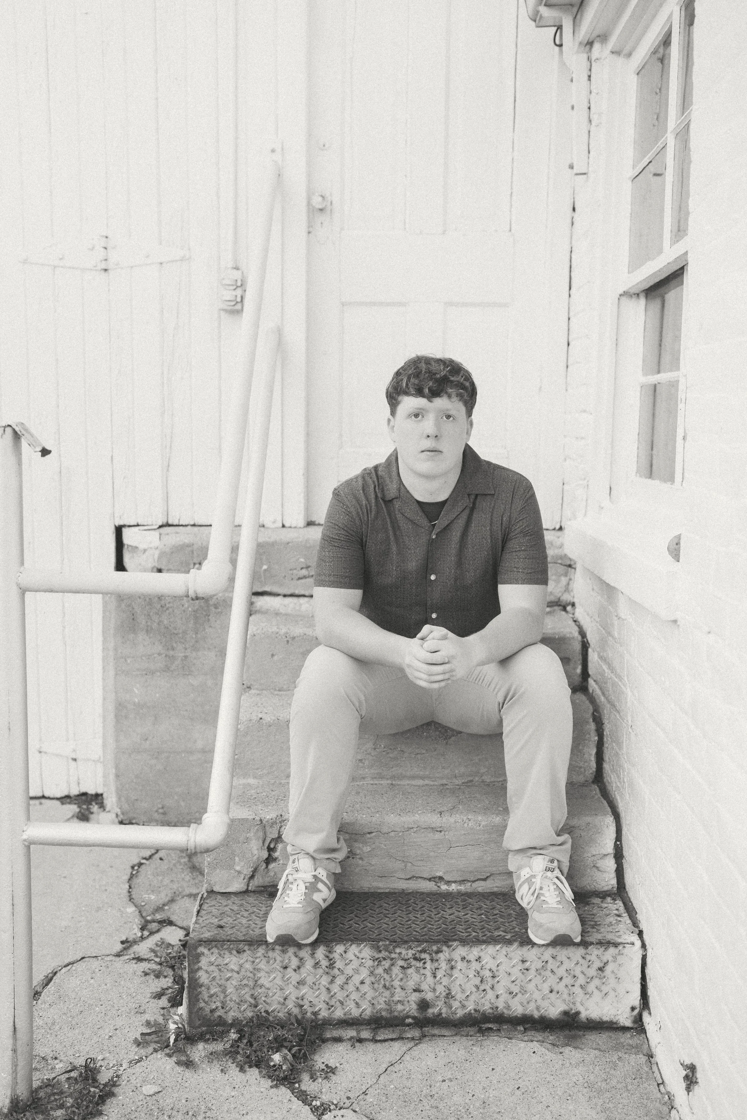 A young man sitting on concrete stairs outside a building, looking directly at the camera, with a serious expression.
