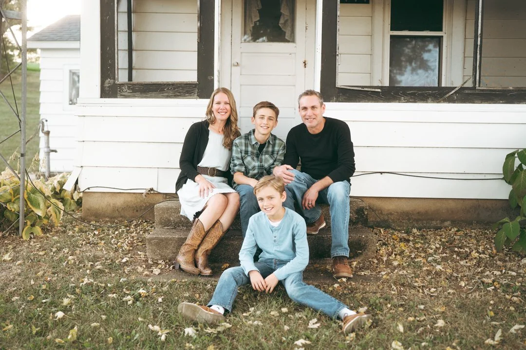 A family of four sitting on the front steps of a white house smiling at the camera, with autumn leaves on the ground.