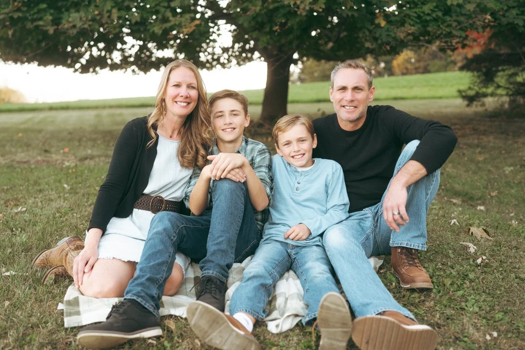 A family of four sitting on a blanket outdoors in a park, smiling at the camera with trees and grass in the background.