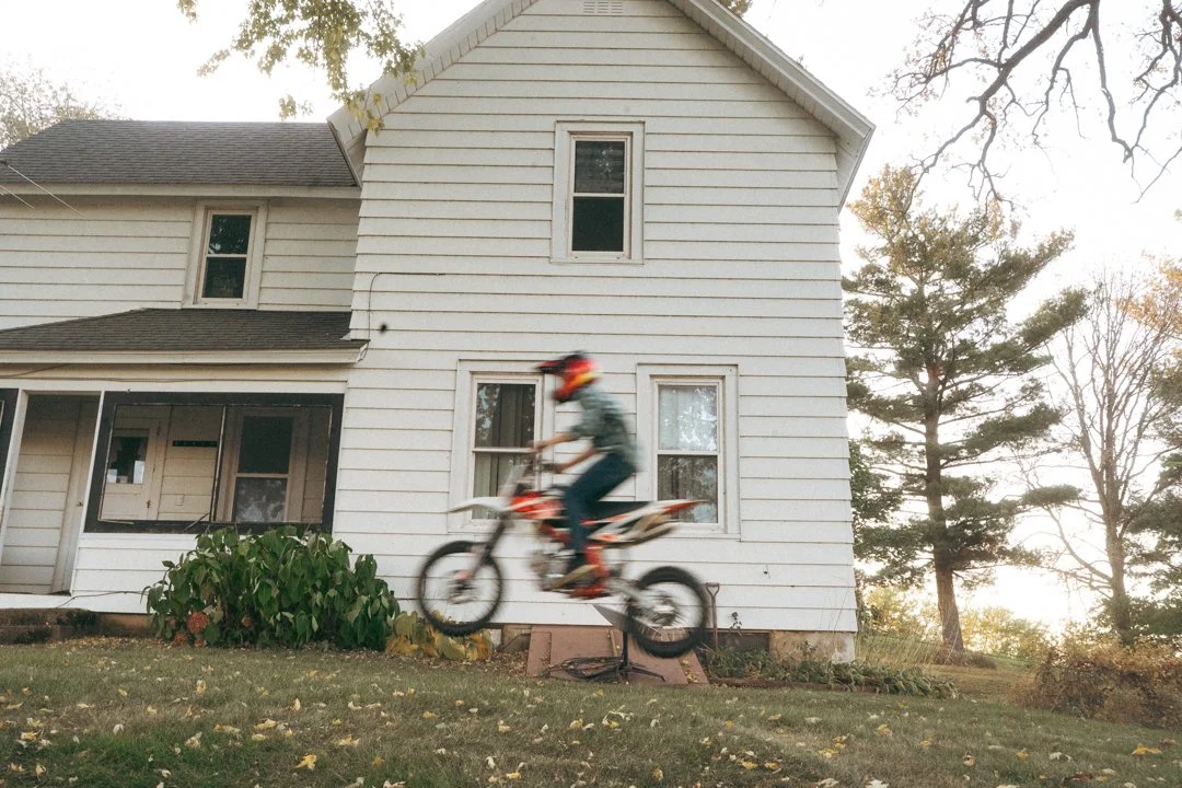 A person wearing a helmet riding a dirt bike in front of a white house with multiple windows, surrounded by trees with autumn leaves in the background.