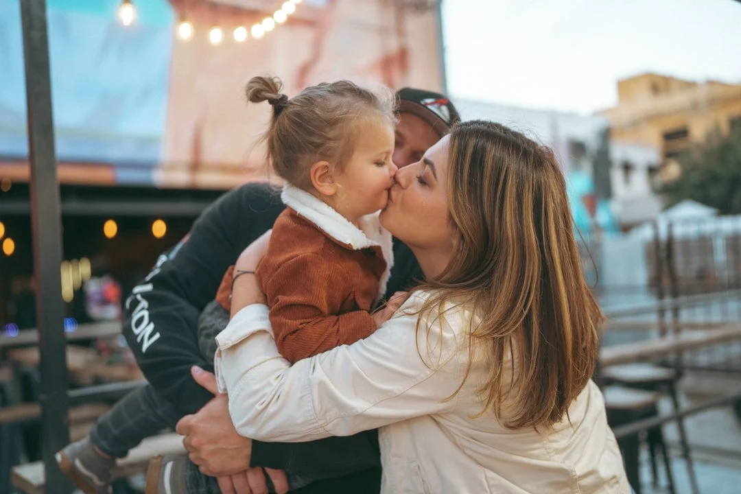 A woman holding a young girl and kissing her on the lips outdoors with blurred background of outdoor seating and string lights.