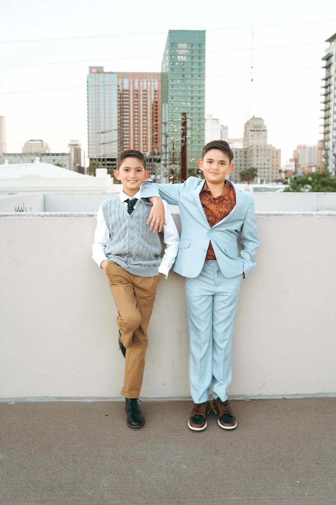 Two young boys posing together on a rooftop with a city skyline in the background, one wearing a light blue suit and the other wearing a gray sweater vest and tan pants.
