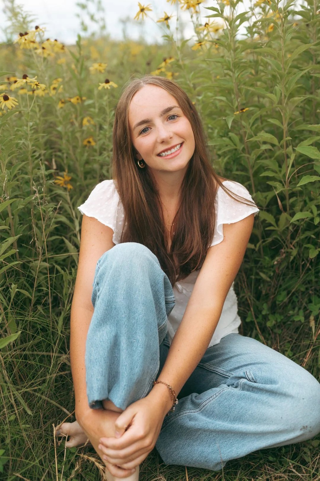 A young woman with long brown hair, smiling, sitting cross-legged in a field of yellow flowers, wearing a white top and jeans.