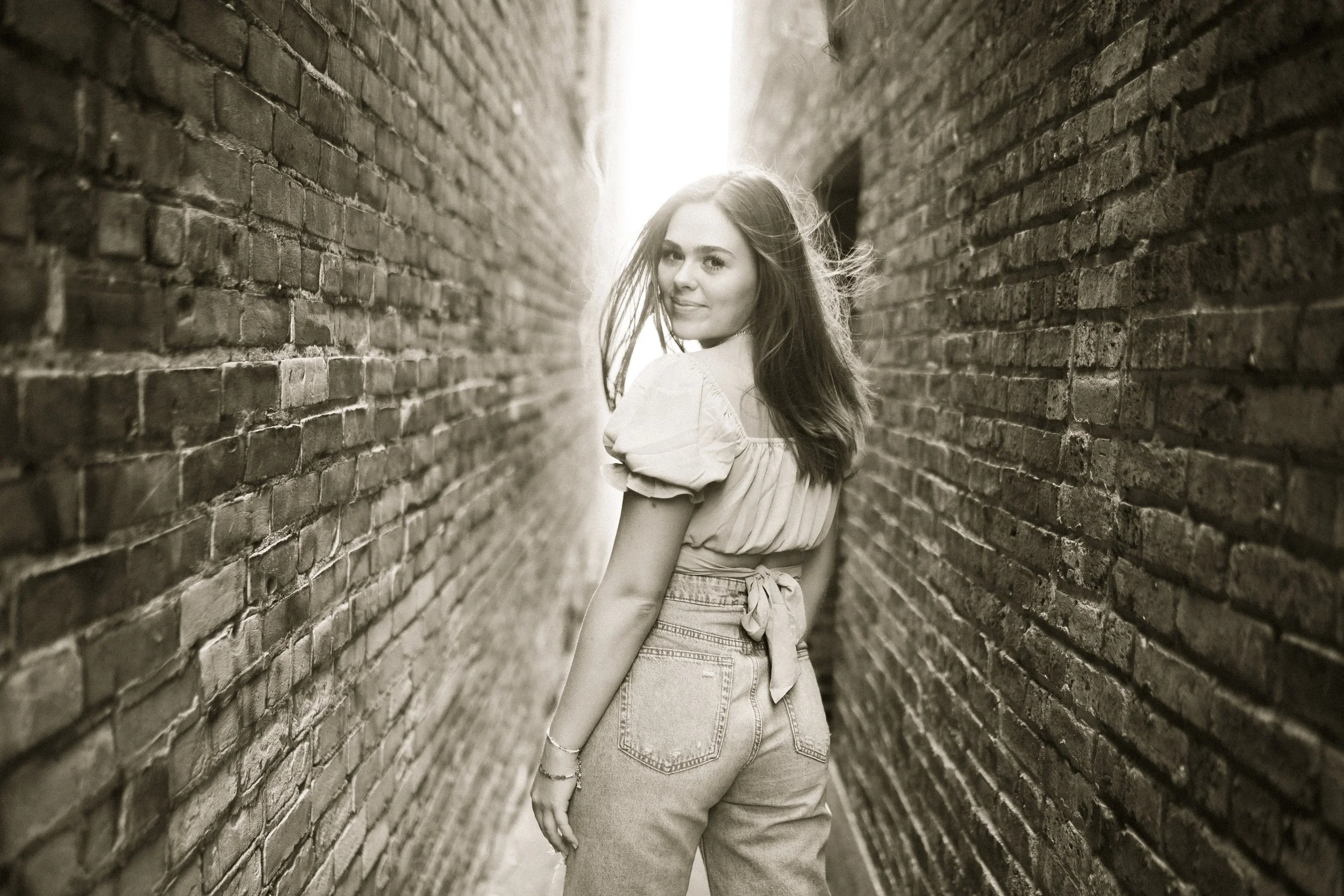 A young woman with long hair smiling and looking over her shoulder, standing between two brick walls with sunlight behind her.