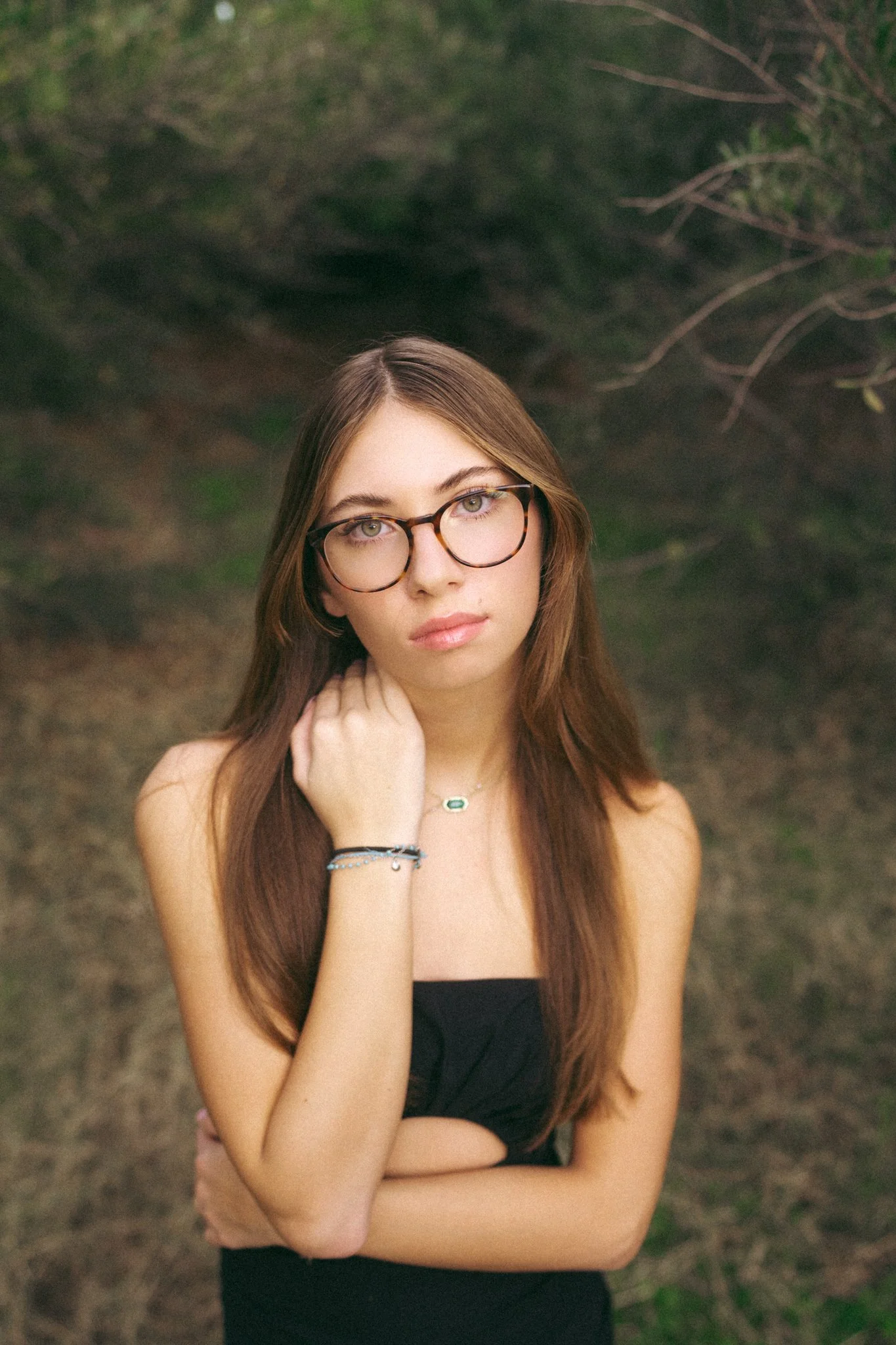 A young woman with long brown hair and glasses standing outdoors with trees and greenery behind her.