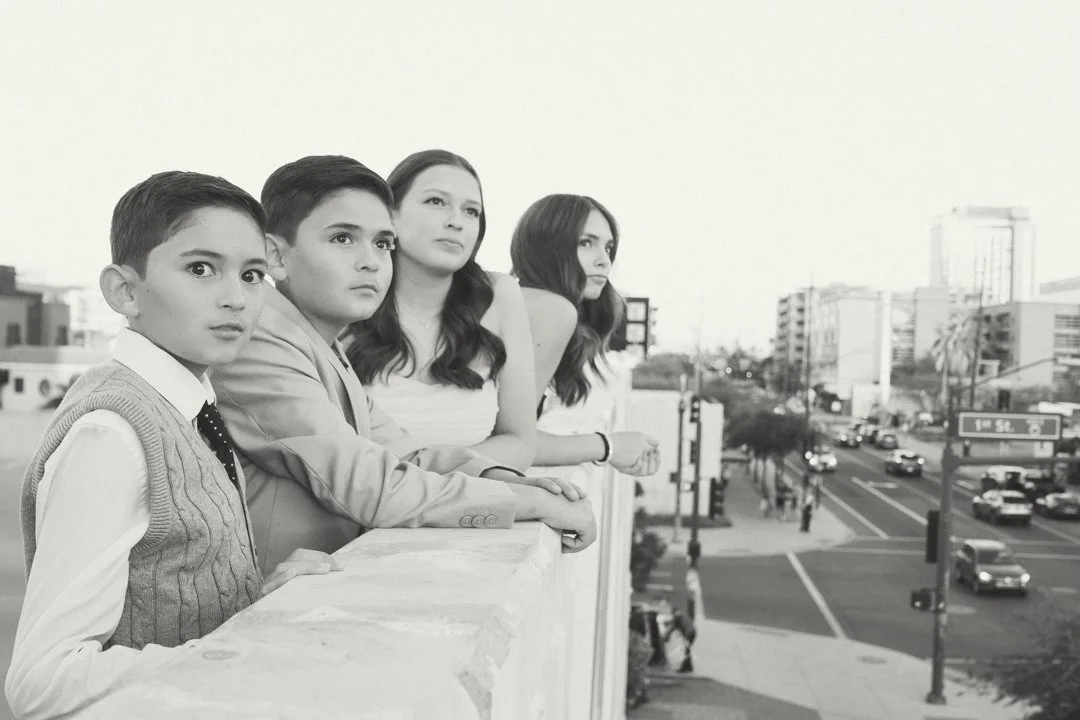 Four young people, three females and one male, standing on a rooftop overlooking a city street, engaged in conversation.