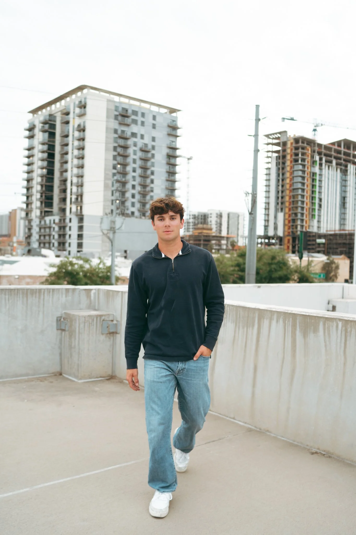 Young man in a black long-sleeve shirt and blue jeans walking on a parking garage rooftop with buildings and cranes in the background.