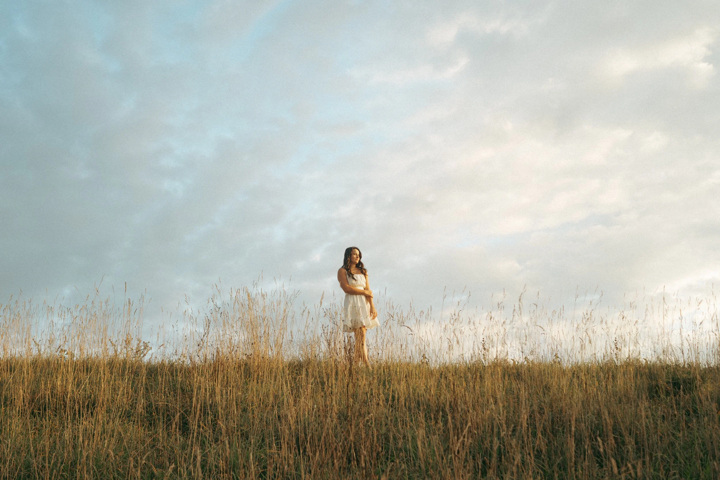 A woman in a white dress standing alone in a grassy field during sunset or sunrise.
