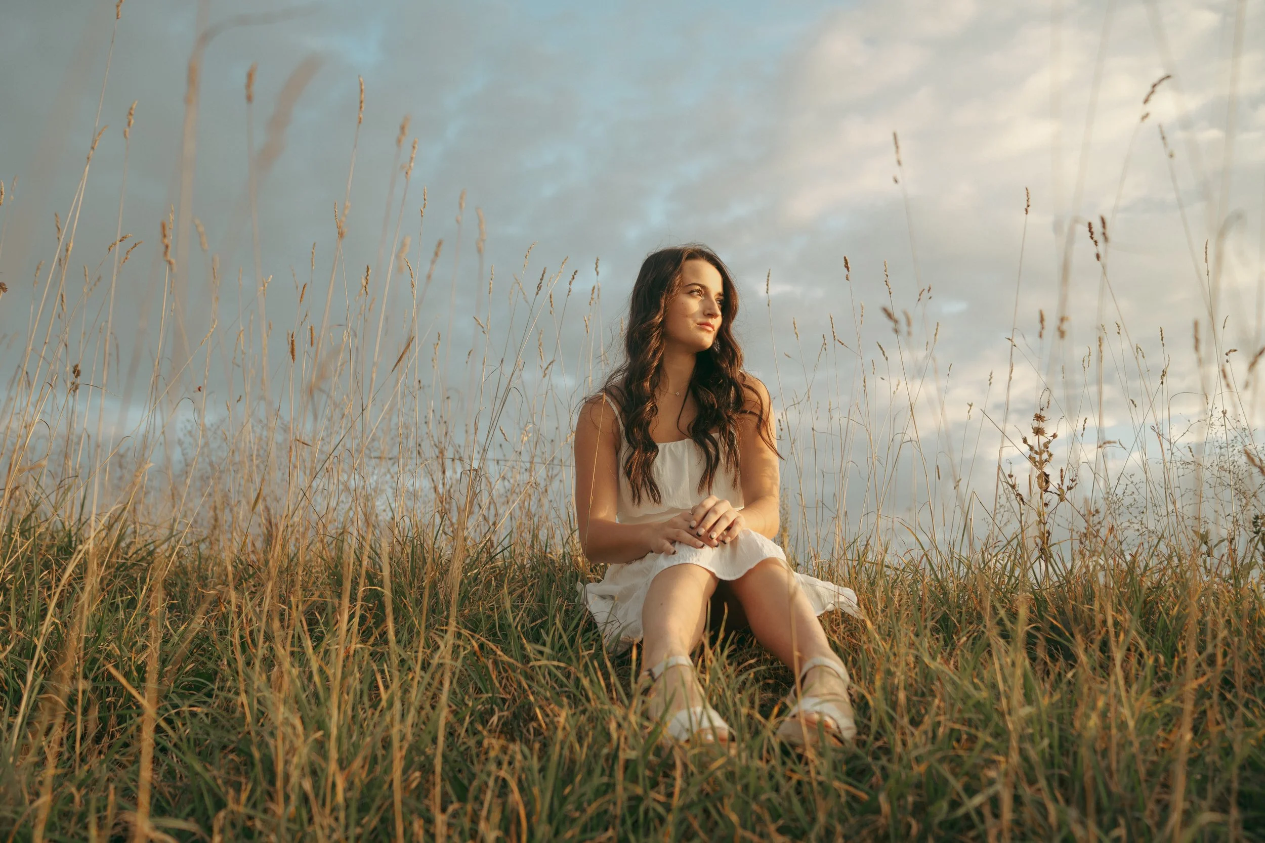 A young woman with long dark hair sitting in a field of tall grass, looking into the distance during sunset.