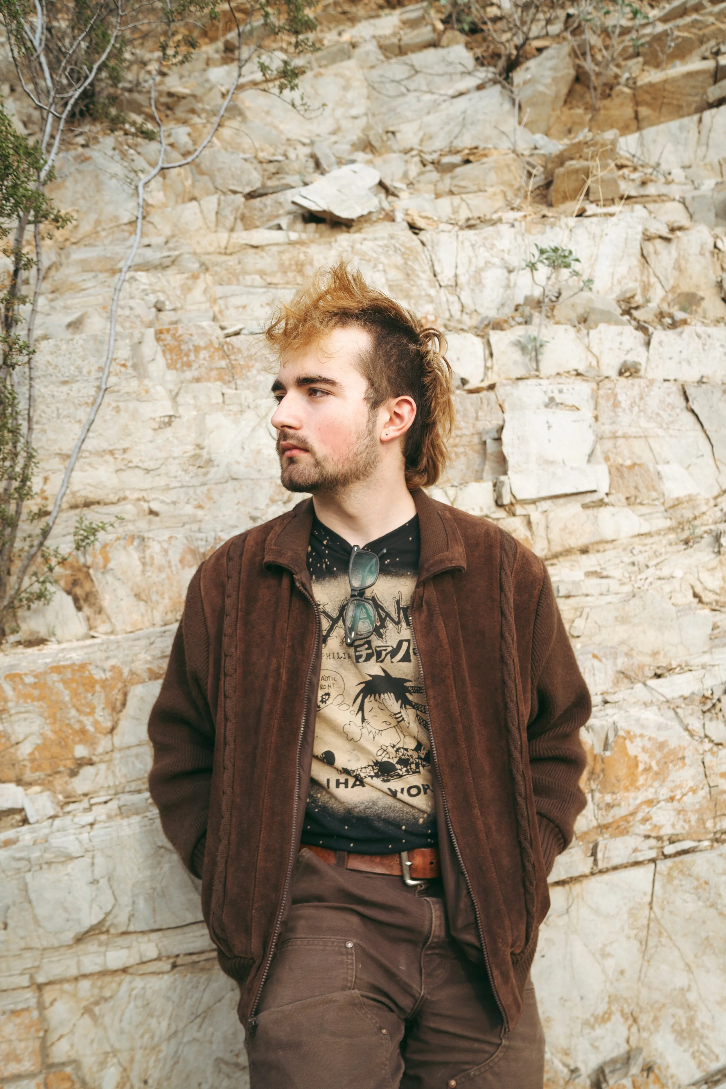 A young man with a mullet hairstyle wearing a brown corduroy jacket, graphic T-shirt, and brown pants, standing against a rocky beige and cream-colored stone wall, looking to his left.