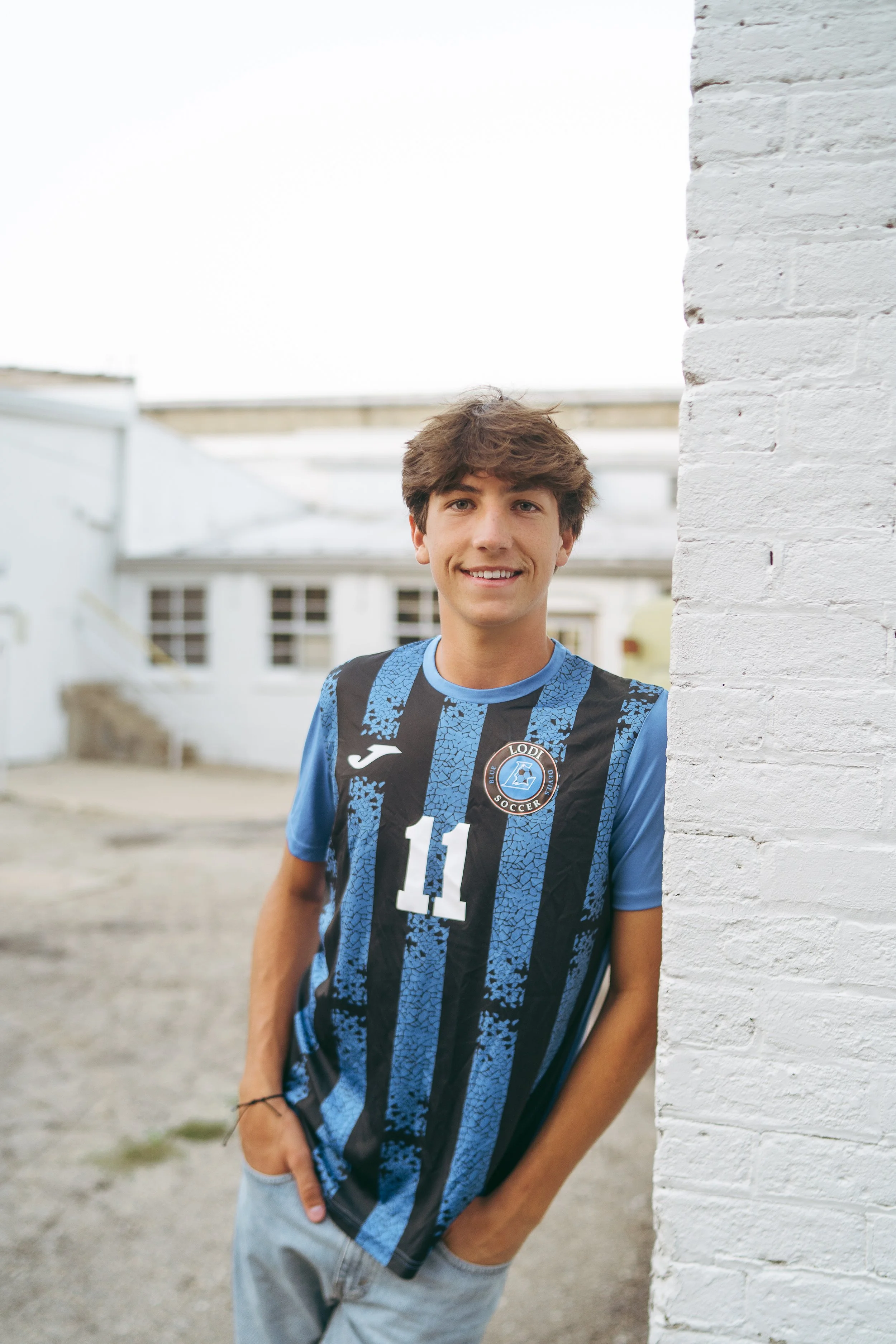A young man with brown, tousled hair is leaning against a white brick wall, smiling at the camera. He is wearing a blue and black soccer jersey with the number 11 and a logo on the chest, along with light-colored pants.